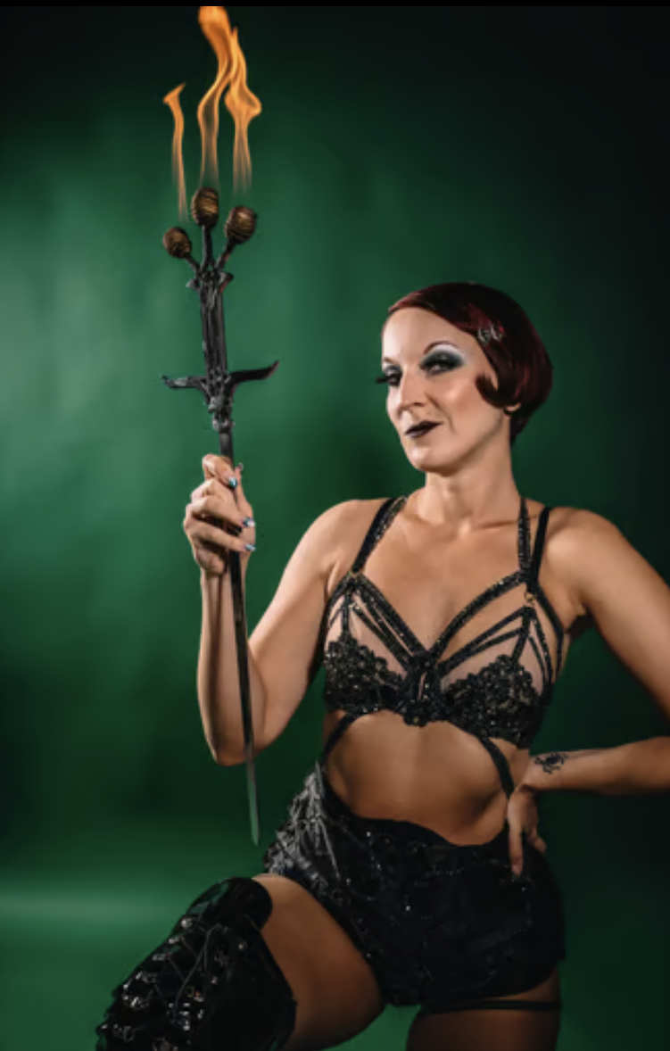 Woman with short red hair, dark makeup, and black lace lingerie holding a flaming trident against a green background.