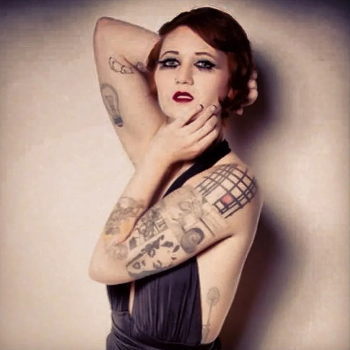 A woman with short red hair and tattoos on her arm and shoulder, posing with one hand on her chin and the other behind her head, wearing a dark fringed top.