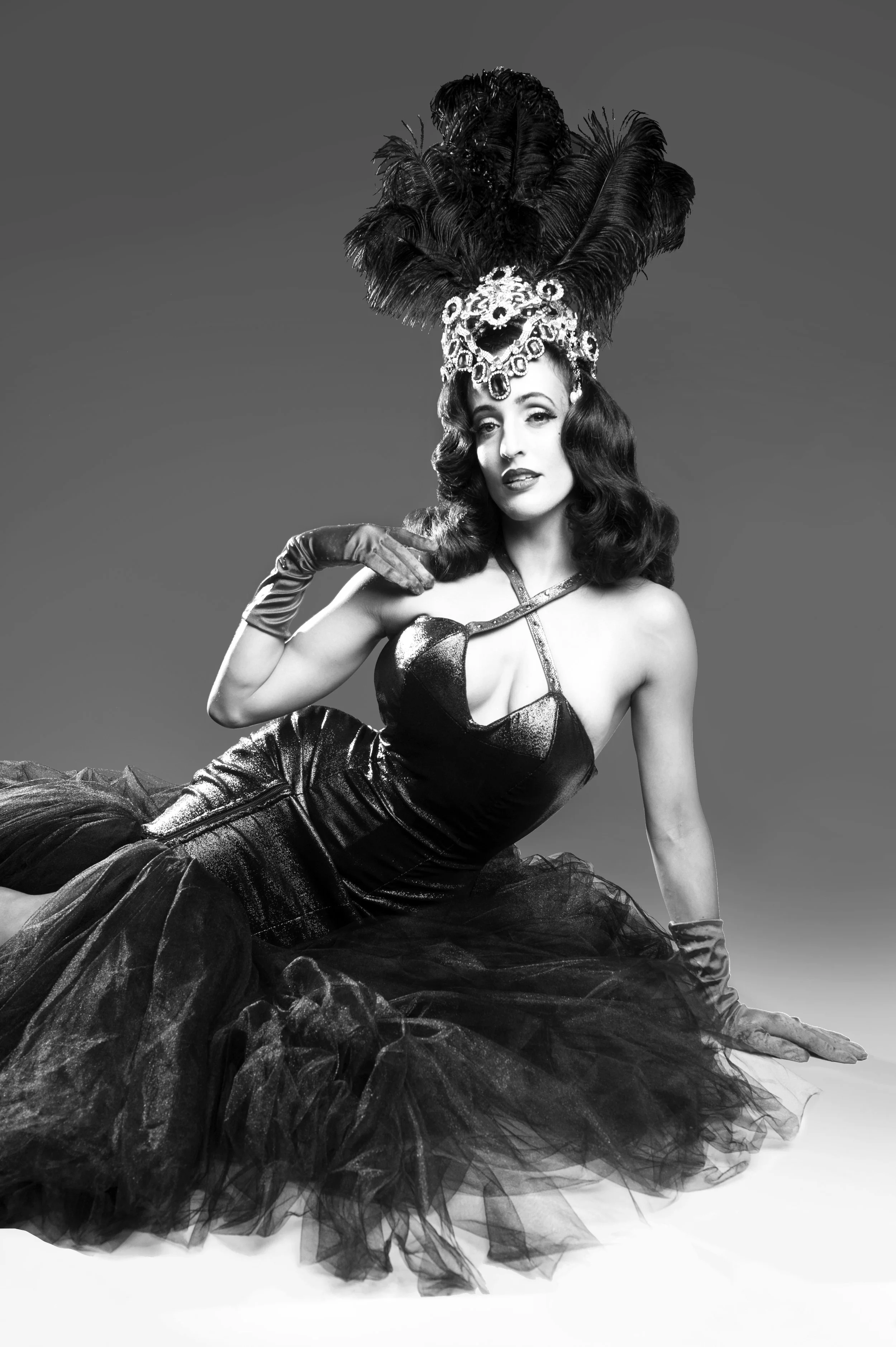A woman dressed in vintage glamour attire, wearing an ornate feathered headdress and a strapless gown, sitting on the ground with one hand resting on her knee, in a black and white studio photograph.