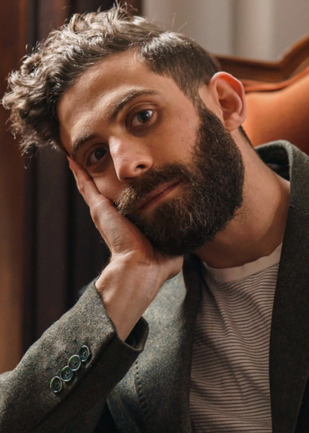 A man with a beard and curly hair resting his head on his hand, sitting in a wooden chair indoors, looking at the camera with a neutral expression.