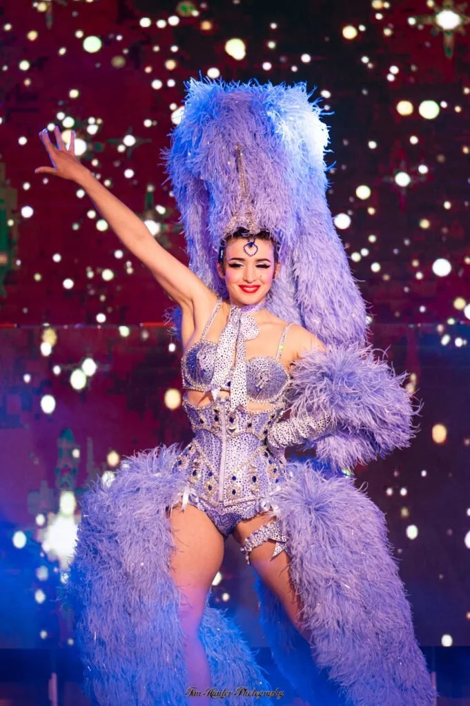 A performer in a purple feathered costume on stage, with one arm raised and smiling, against a festive, sparkling background.