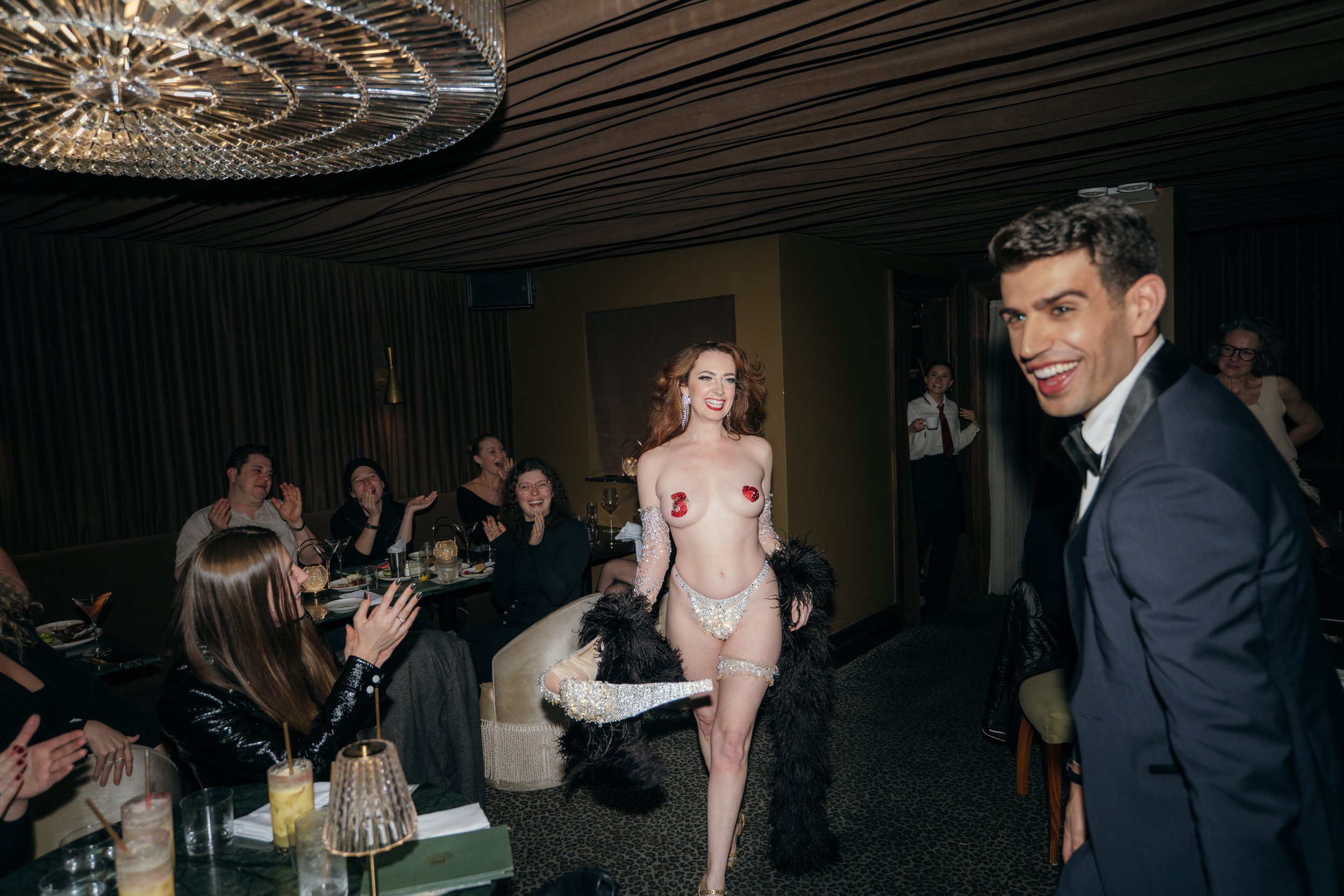A woman wearing revealing silver lingerie with the number 3 and a heart made of red fabric covers on her nipples, walking in a room full of smiling and clapping people at a party. She has long, curly red hair and is holding a black feathered boa. A man in a tuxedo is looking at her, smiling.