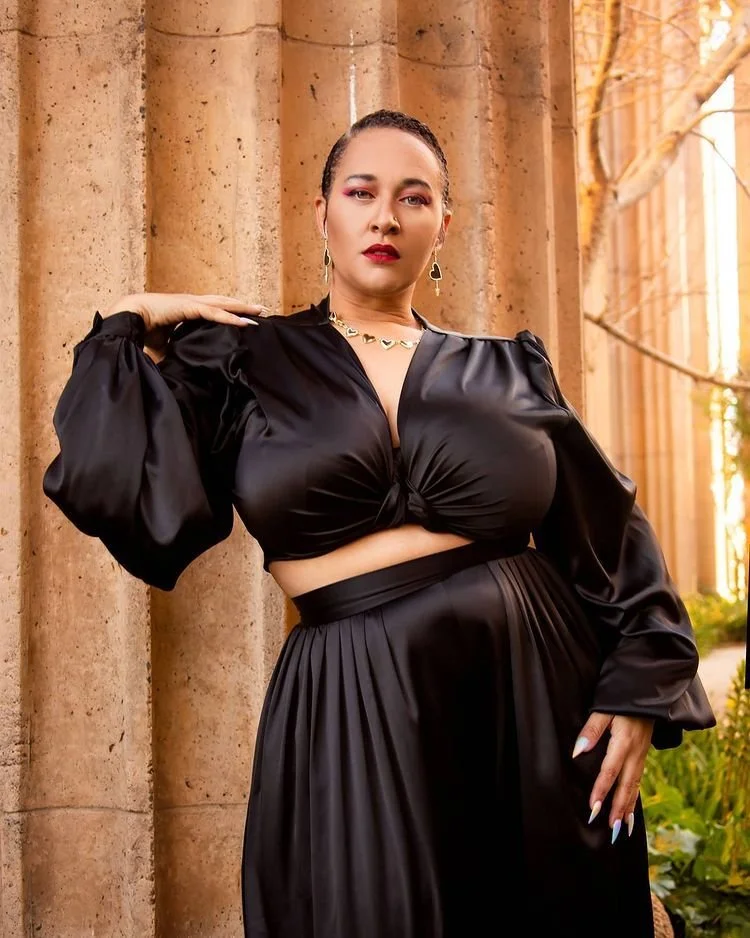 A woman in a black satin outfit posing outdoors in front of stone columns, with sunset lighting and trees in the background.