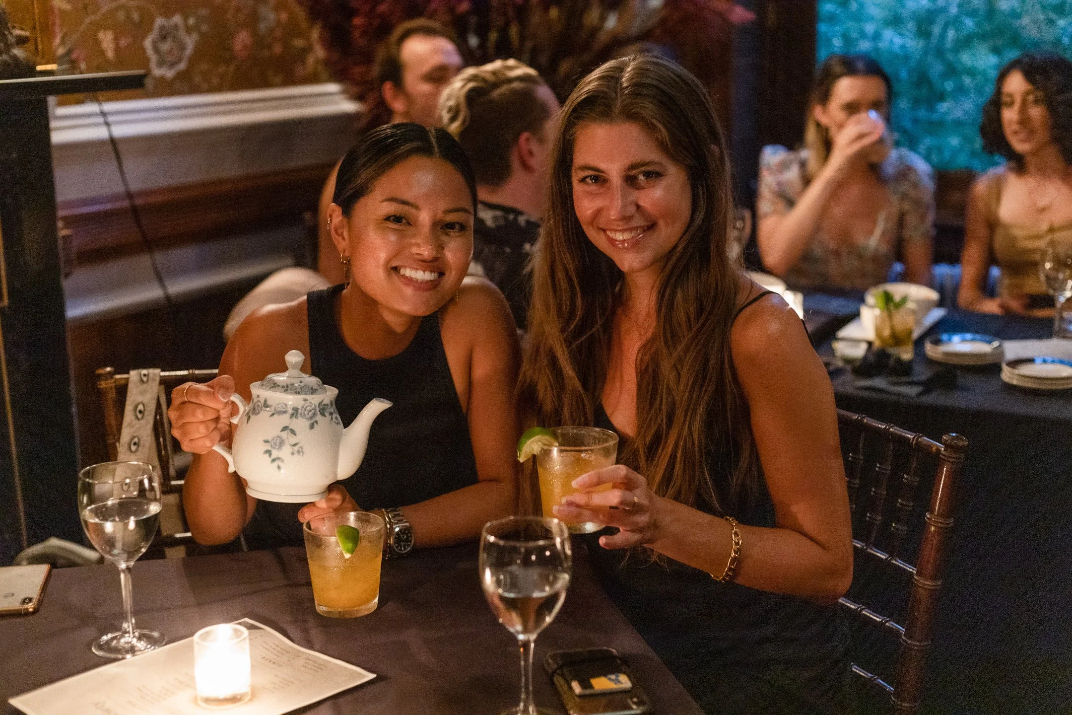 Two women smiling at a restaurant table, one holding a teapot and the other holding a cocktail with lime, surrounded by glasses, a candle, and table settings.