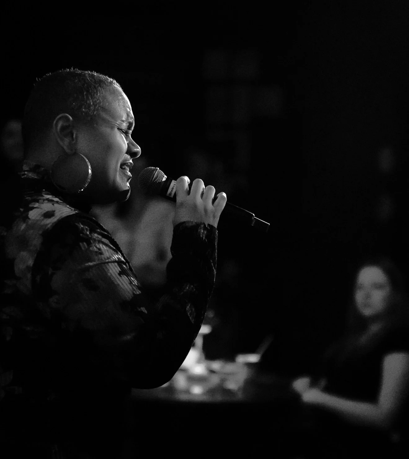 A woman singing into a microphone with her eyes closed, wearing large hoop earrings and a patterned top, in a dark setting.