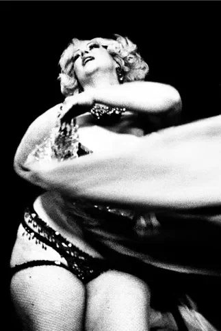A performer with curly blonde hair and revealing attire, possibly a costume or stage outfit, is mid-performance with a dramatic expression.