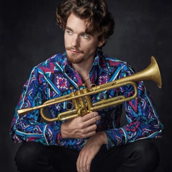 A man with curly brown hair, a beard, and mustache, wearing a colorful patterned shirt, holding a gold trumpet against a dark background.