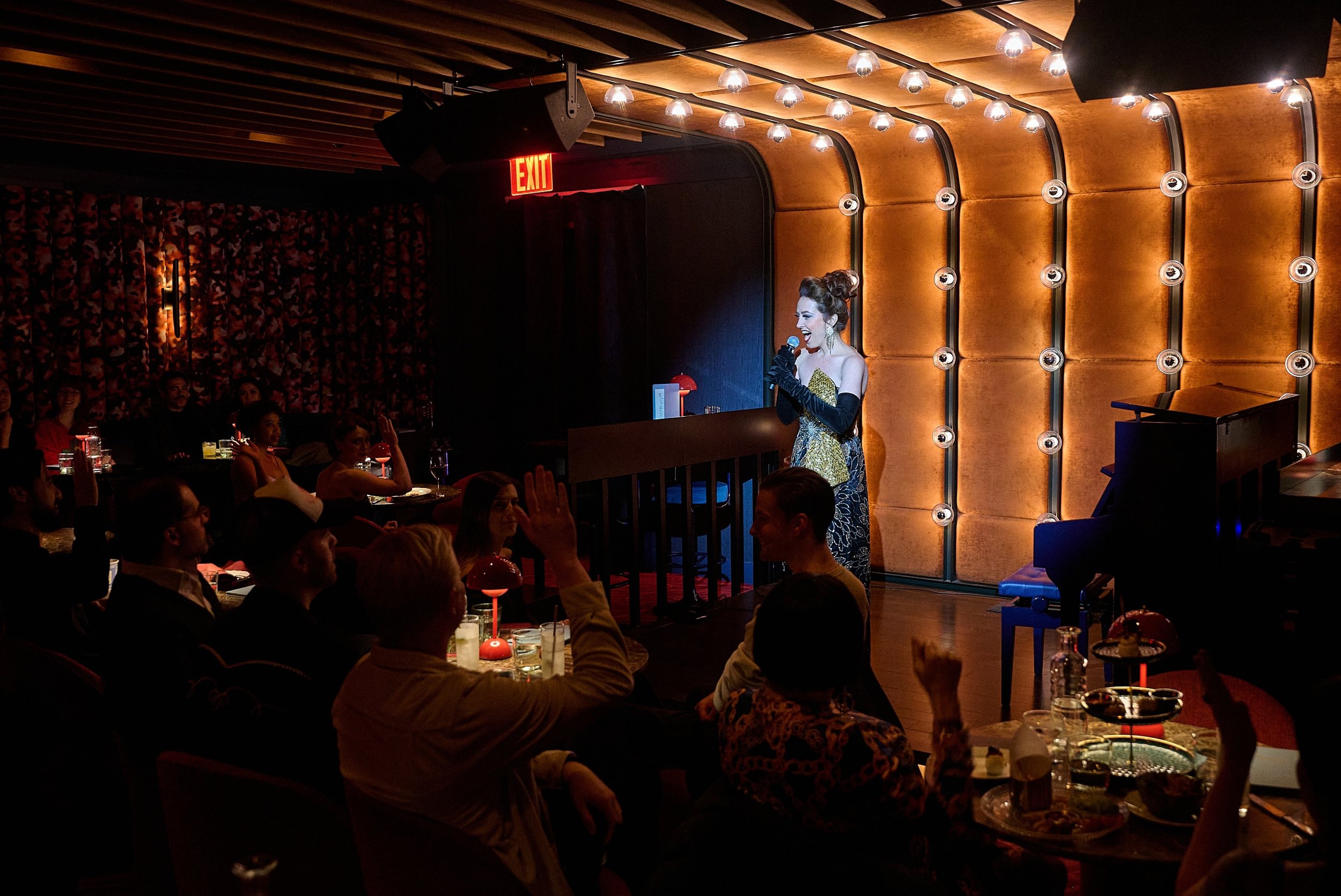 A woman performing on stage at a comedy or musical show in a dimly lit venue with an audience seated at tables, some raising their hands, and warm lighting on the walls.