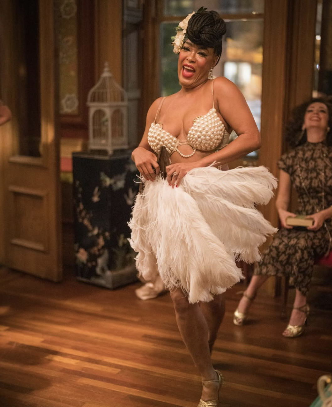 A woman dancing and singing at an event, wearing a pearl-studded bra top and a feathered skirt, with a floral headpiece and earrings, in a warmly lit room.