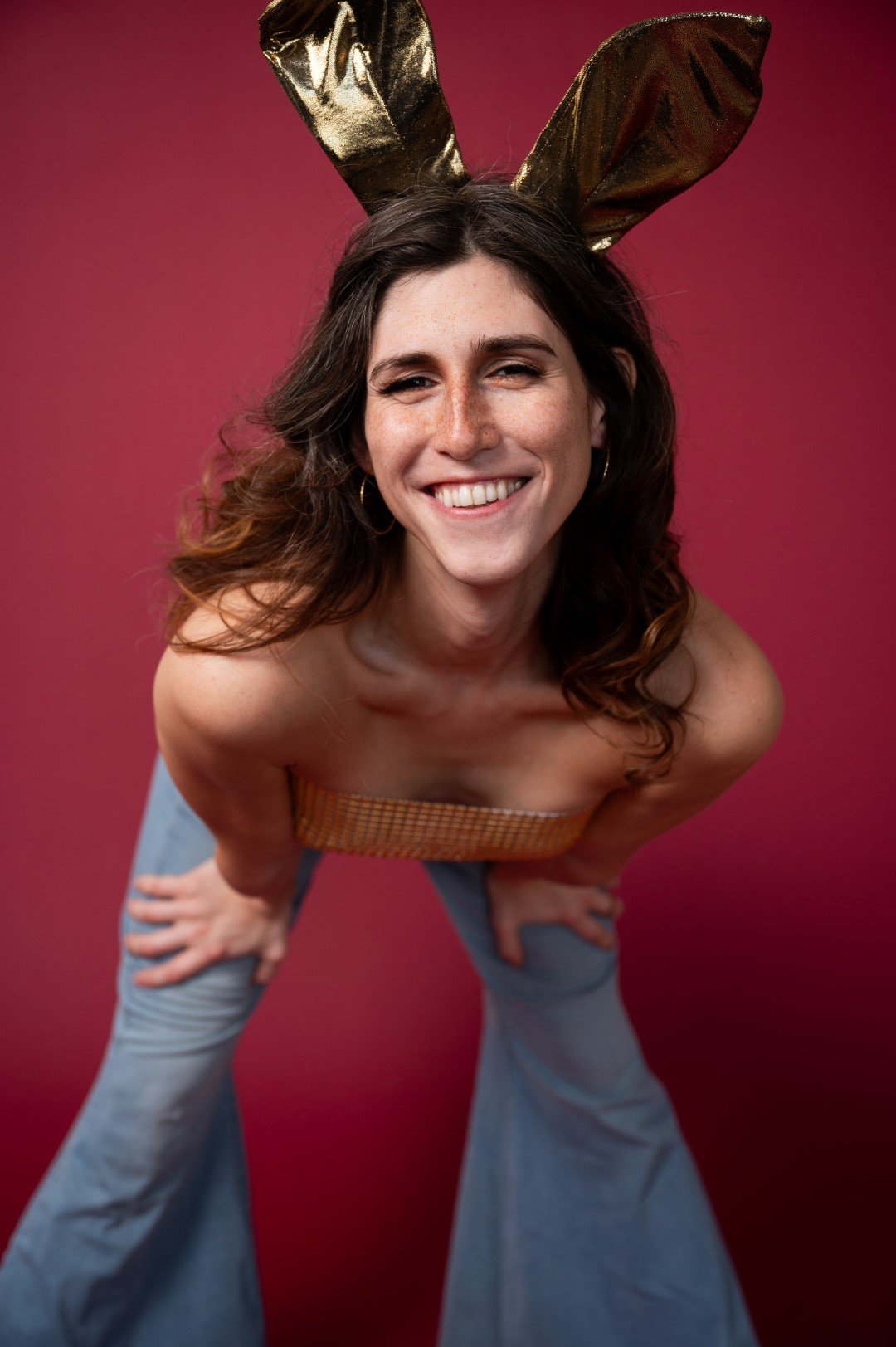 A woman with long wavy brown hair smiling, wearing gold bunny ears, a strapless orange top, and light blue bell-bottom jeans, posing against a pink background.