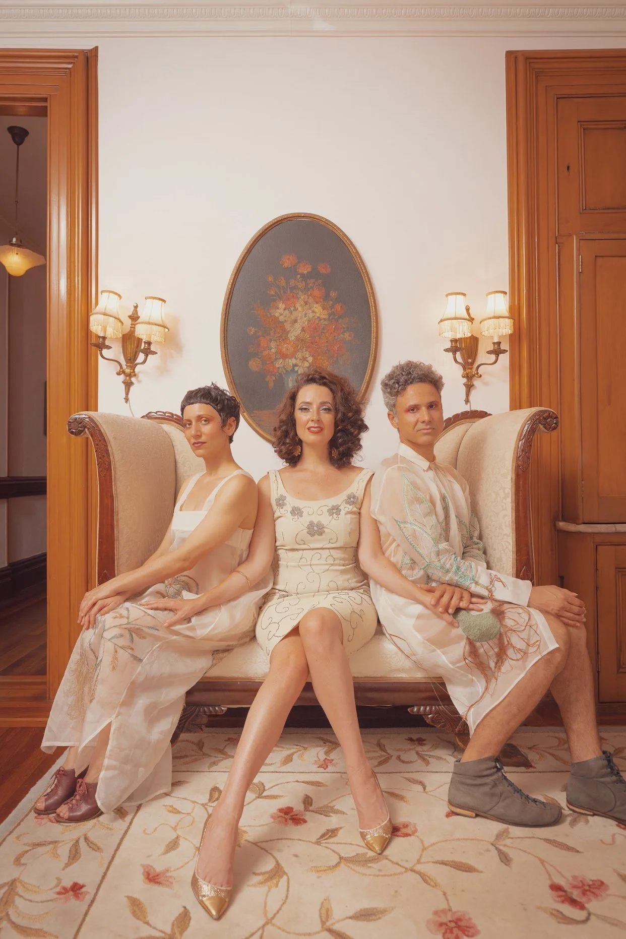 Three people, two women and one man, sitting on a vintage sofa in a living room with wood-paneled walls and ornate lighting fixtures. The woman in the center has curly hair and is wearing a cream dress, while the other woman and man are dressed in similar light-colored clothing with embroidered patterns.
