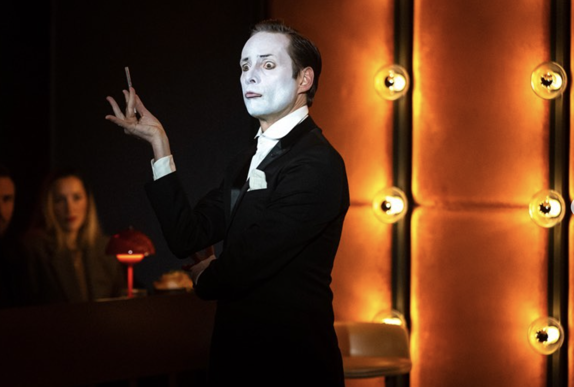 A mime artist dressed in a black tuxedo with white face makeup and a cigarette in hand, standing in a dimly lit performance space with orange glowing wall lights.