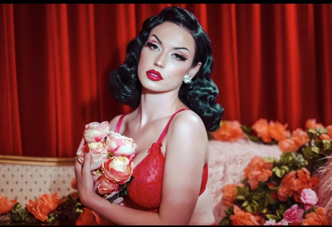 A woman with vintage style black hair, red lipstick, and earrings stands in front of red curtains, holding pink and white roses, surrounded by orange flowers.