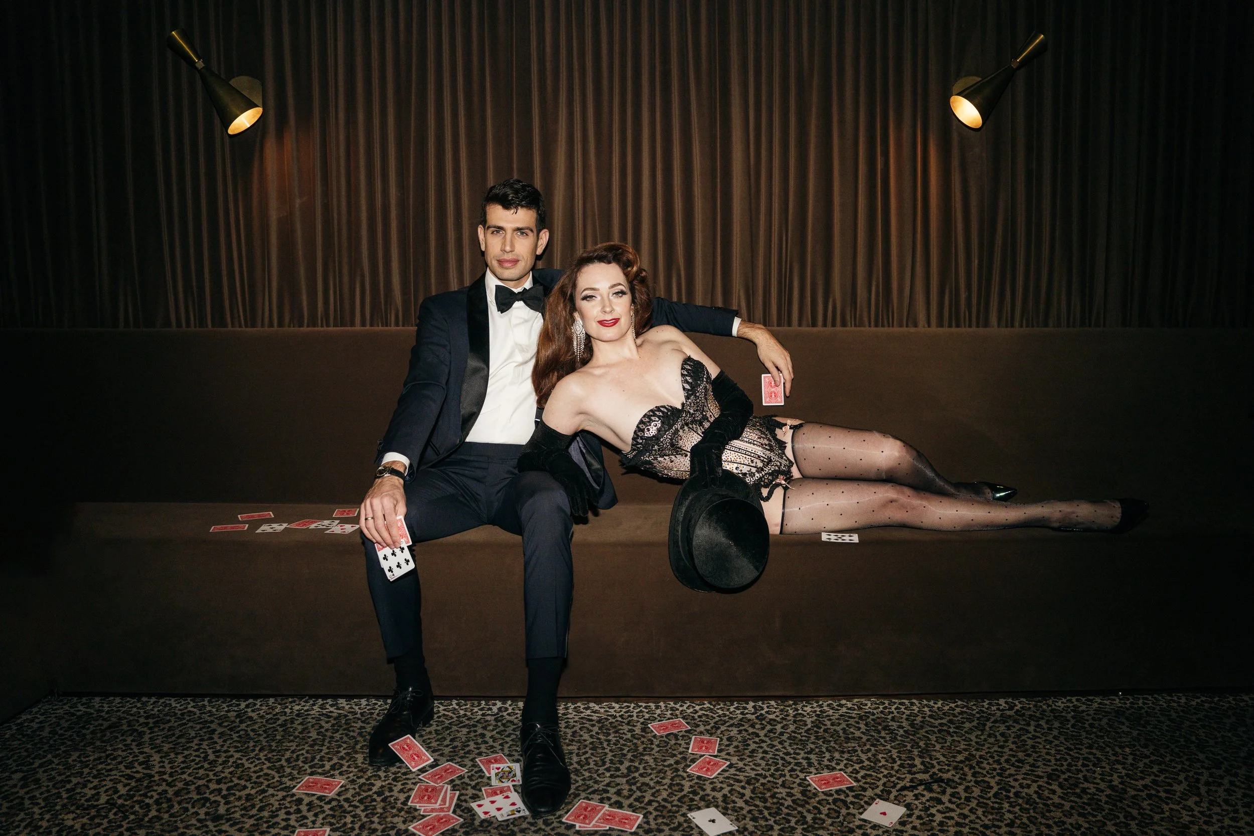 A man and woman dressed in vintage clothing sitting on a bench with playing cards scattered on the floor and bench, with dim lighting and dark curtains in the background.