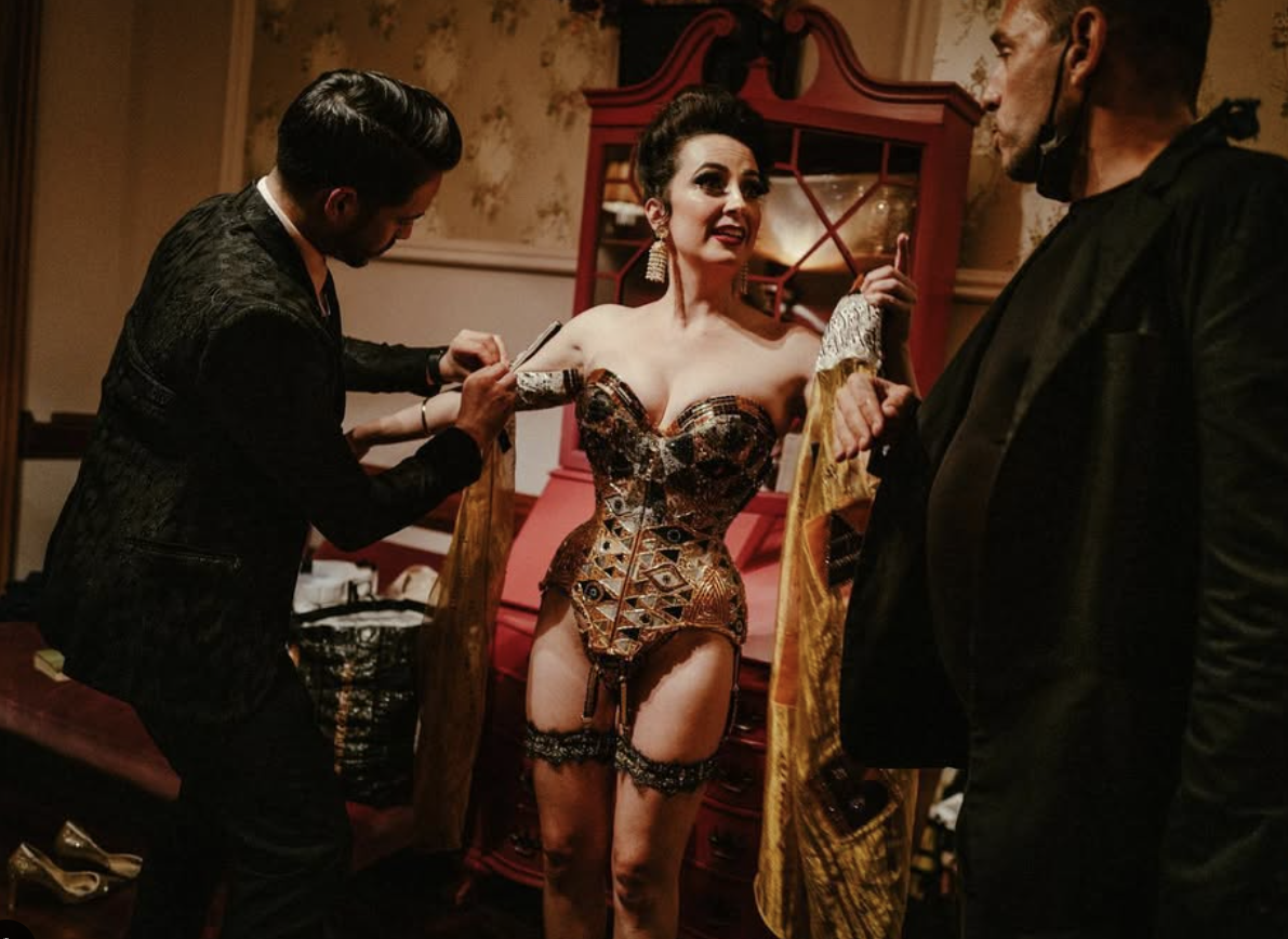 A woman in an ornate corset and stockings stands while two men help her into a jacket in a warmly lit room with floral wallpaper and a vintage red cabinet in the background.