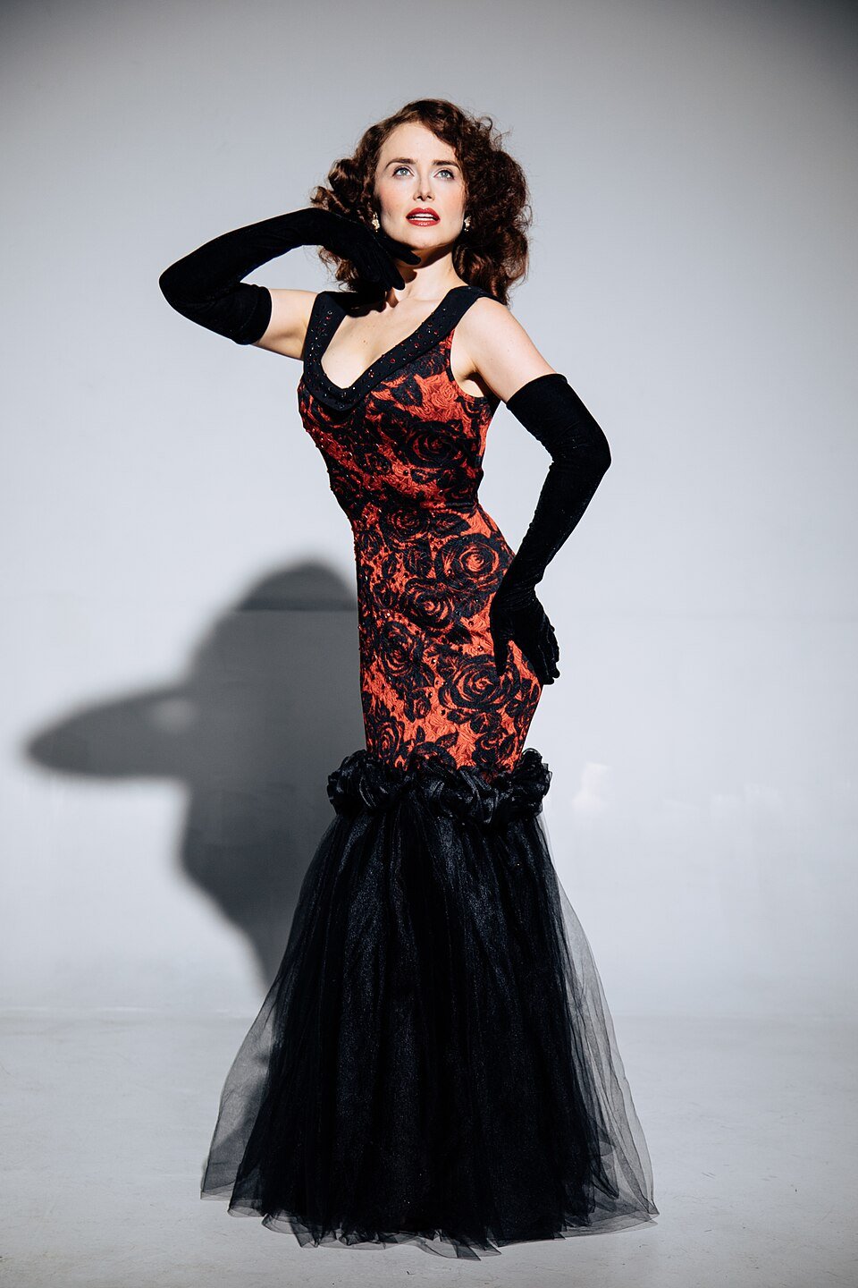 A woman in a red and black lace evening gown with a black tulle train poses with her left hand on her hip and her right hand near her face against a plain white background.