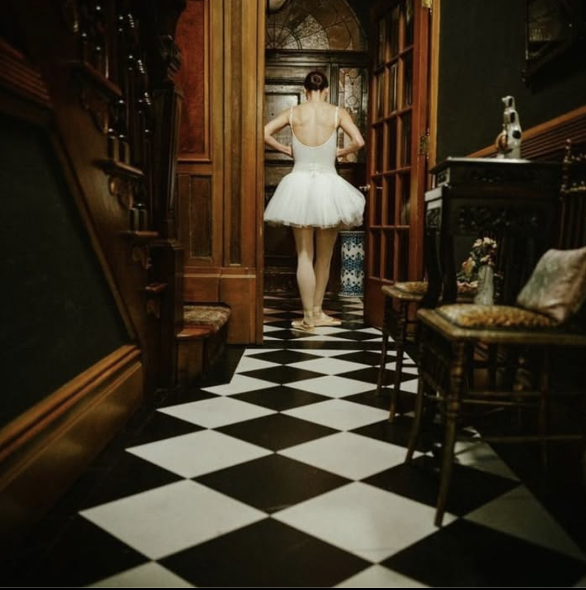 A ballet dancer in a white tutu and tights standing in a vintage room with wooden furniture and black-and-white checkered flooring, seen from behind.