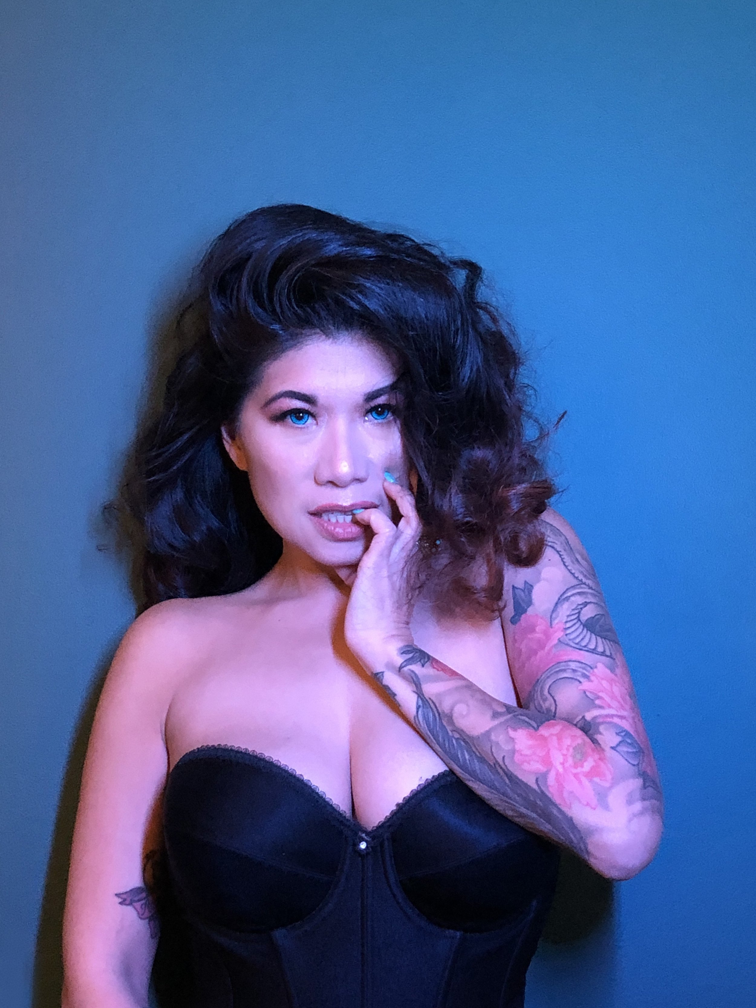 A woman with long dark curly hair and bright blue eyes poses against a plain background. She is wearing a black strapless top and has colorful tattoos on her arms, with one visible on her left arm.