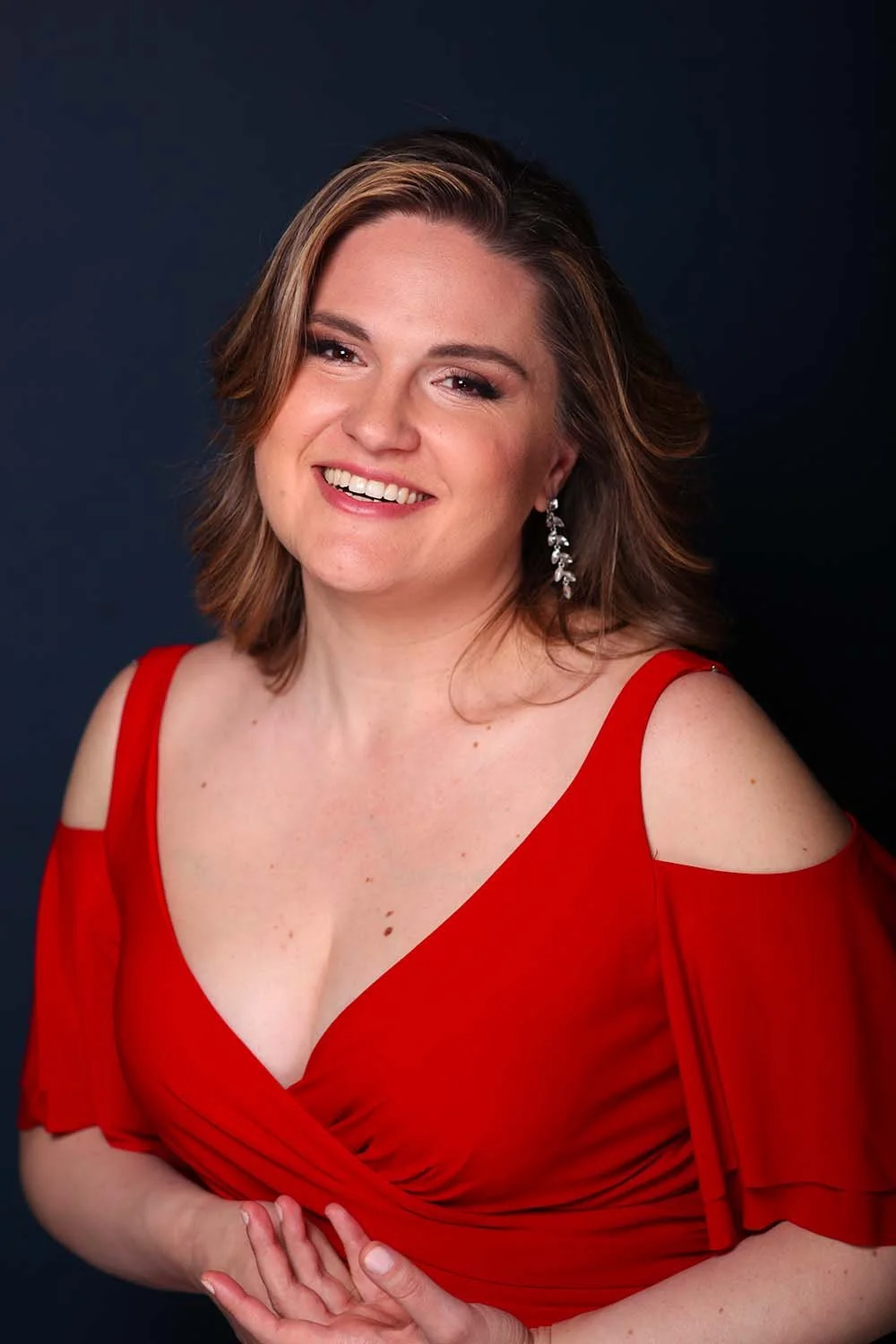 A woman with shoulder-length brown hair wearing a red dress with cut-out shoulders and sparkling earrings, smiling against a dark background.