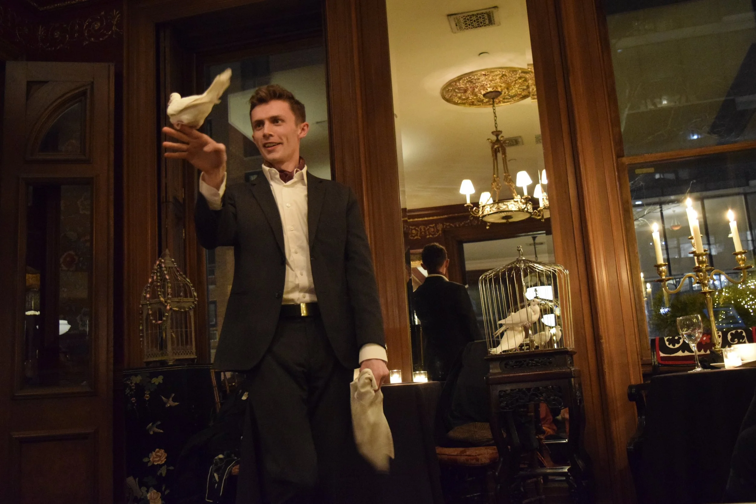 A waiter in a tuxedo serving food or drink in an elegant restaurant with wood-paneled walls, chandeliers, and candlelit tables.