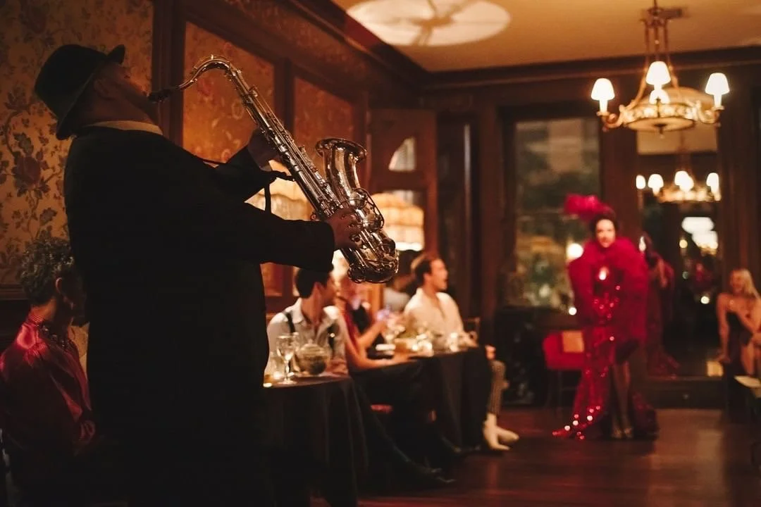 A jazz musician playing the saxophone at an elegant indoor event with seated guests and a singer in a red dress in the background.