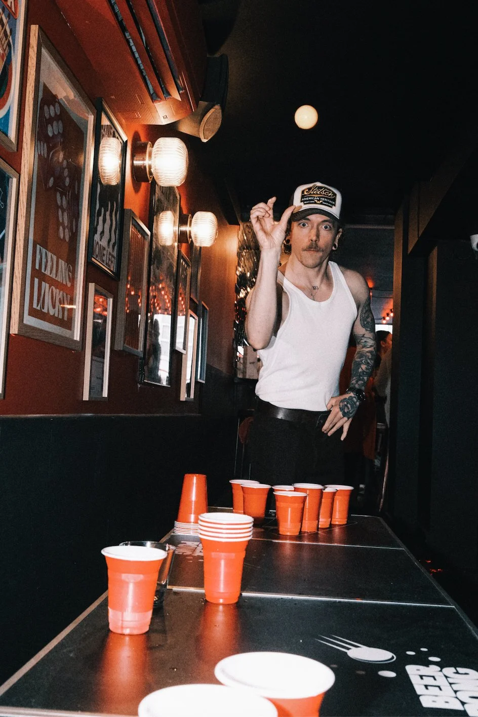 A man with tattoos, wearing a white tank top and a trucker hat, stands in a dimly lit bar or party setting, preparing to toss a beer pong ball at a table with red cups arranged for a game.