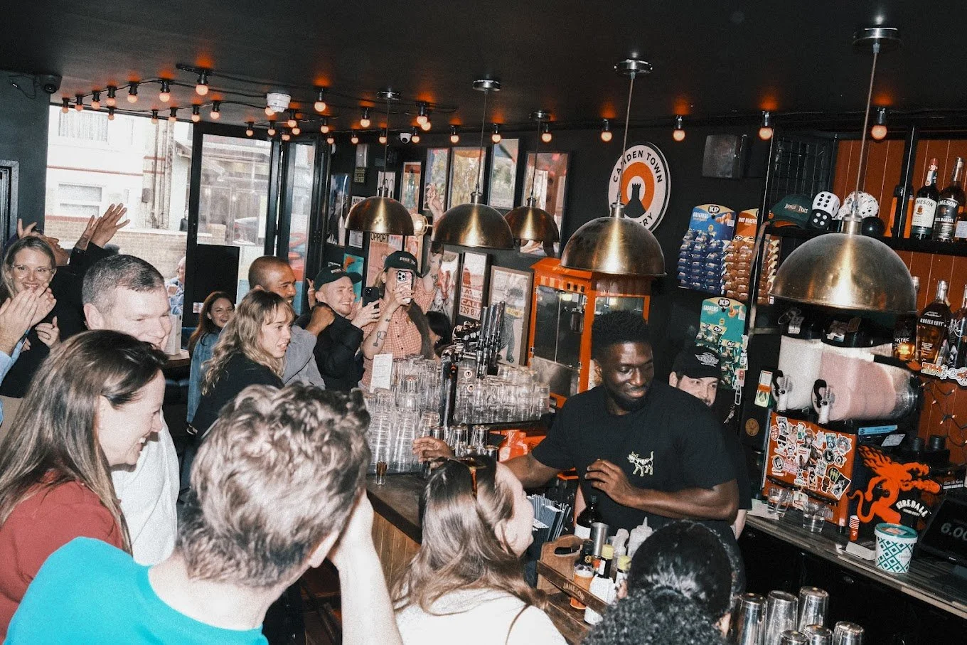 A lively bar scene with a bartender preparing drinks, surrounded by an engaged crowd of patrons in casual attire, with some taking photos and others watching, inside a trendy bar decorated with warm lighting and eclectic decor.