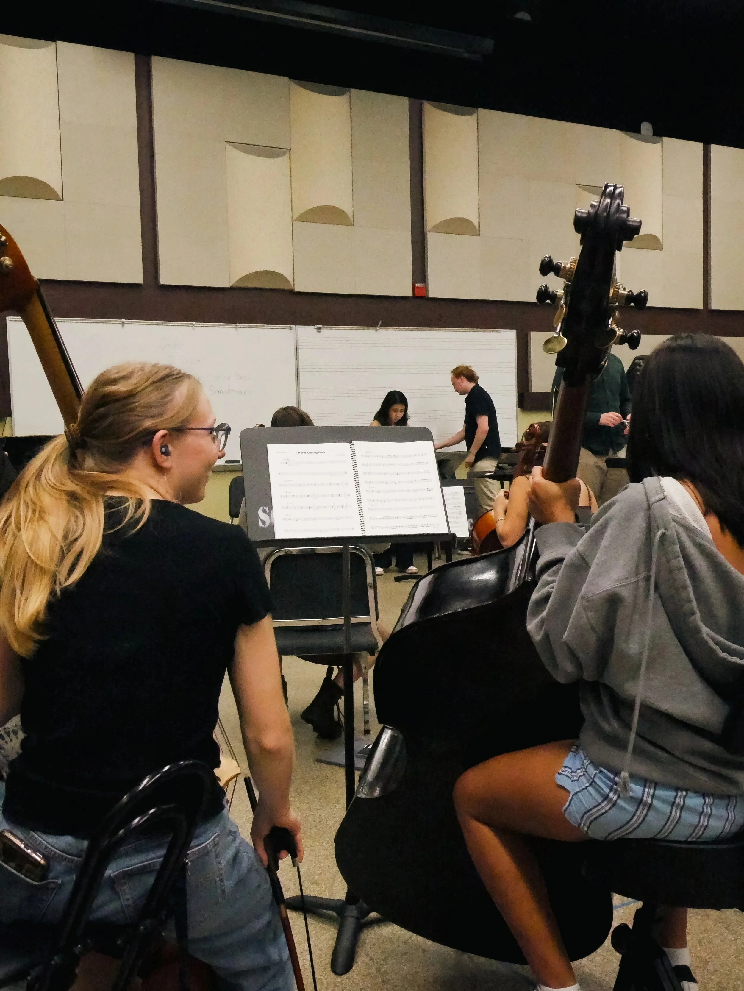 A group of young students in a music classroom is preparing for a rehearsal. Visible are a girl with glasses and blonde hair, people with musical instruments including a violin and a double bass, and a whiteboard with musical notes and writing.