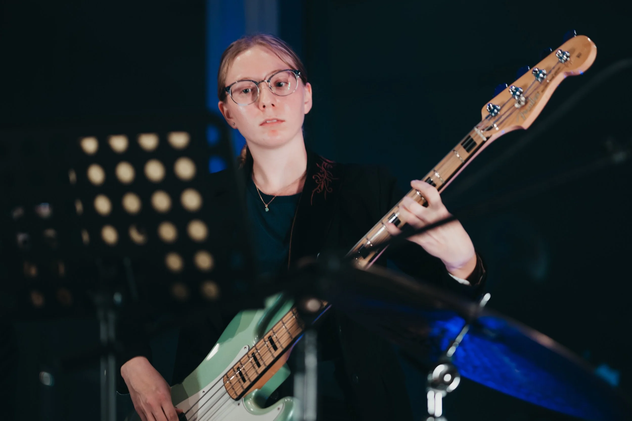 A person wearing glasses playing an electric guitar on stage, with music stands and stage lights in the background.