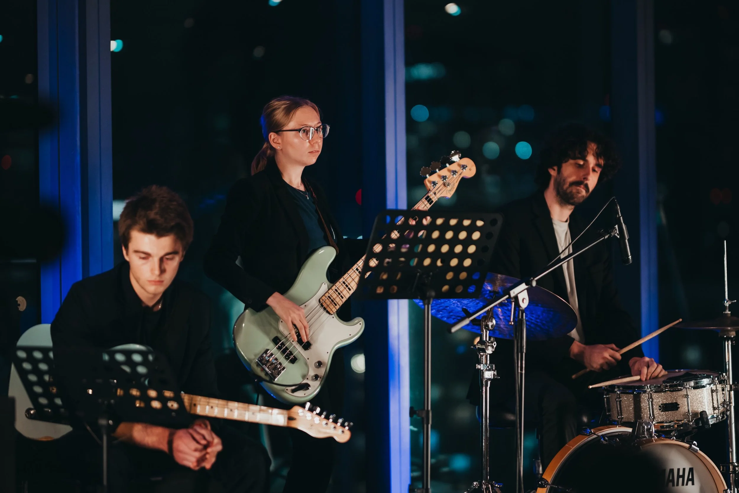 A group of musicians performing at a night event, including a woman with glasses playing an electric bass guitar, a young man playing an acoustic guitar, and another man playing drums, all seated near large windows with city lights visible outside.
