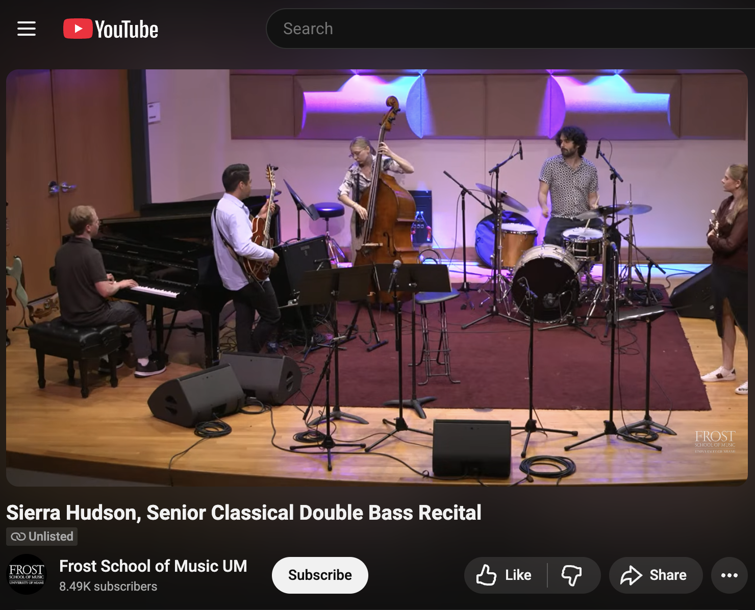 A group of five young musicians performing on stage with various instruments: a pianist, a guitarist, a double bassist, a drummer, and a vocalist. The stage has a purple and blue lighting setup and is set for a musical recital.