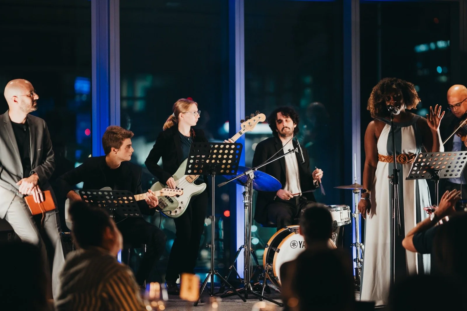 A live band performs on stage indoors at night, with city lights visible through large windows behind them. The band consists of six musicians: a man in a blazer on the left, a seated guitarist, a female bassist, a drummer, a female singer with a mic