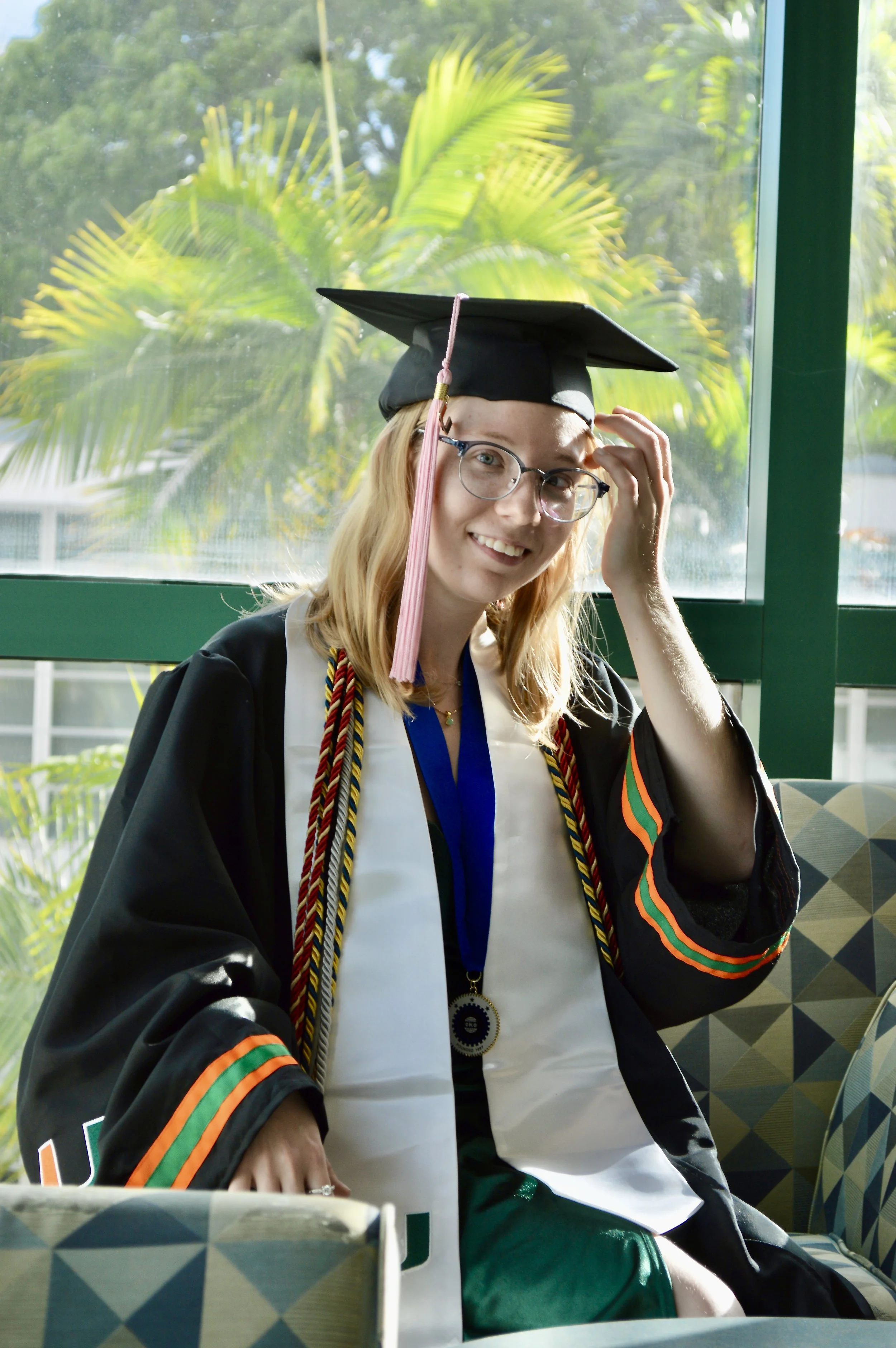 A woman wearing a black graduation gown and cap, glasses, and a diploma while sitting indoors with a green outdoor background visible through windows.