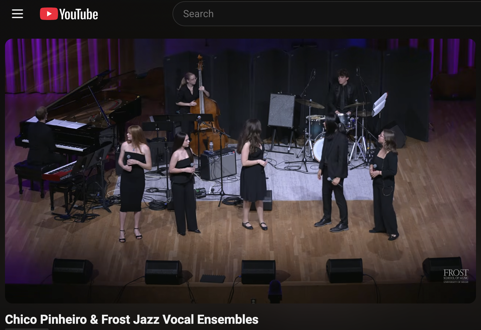 A jazz vocal ensemble performing on a stage, with six singers holding microphones, accompanied by a pianist, double bassist, and drummer. The stage is set in a concert hall with purple curtains and a hardwood floor.