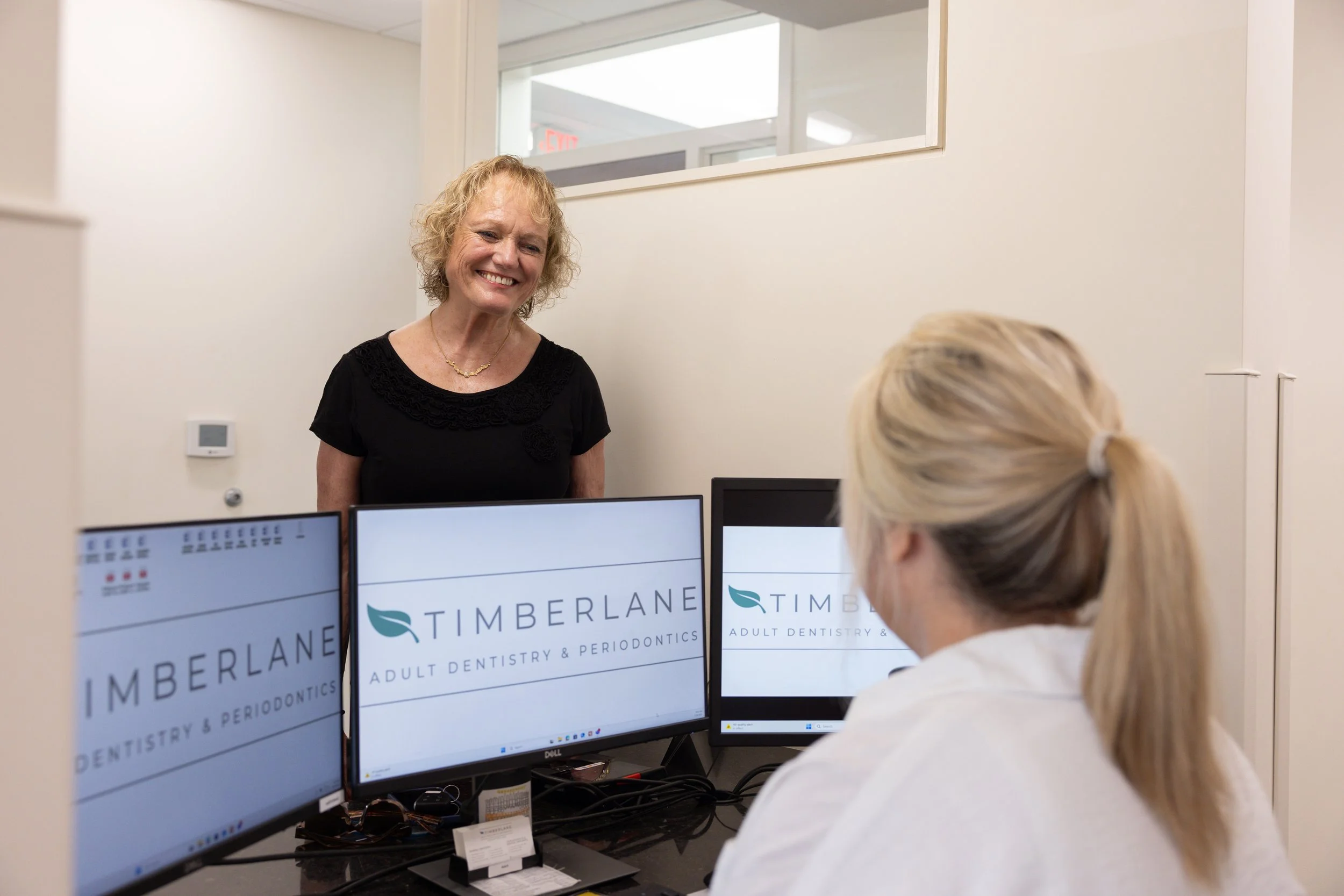 A woman with curly blonde hair smiling at a reception desk with computer monitors displaying 'TIMBERLANE Adult Dentistry & Periodontics' signage, inside a dental office.