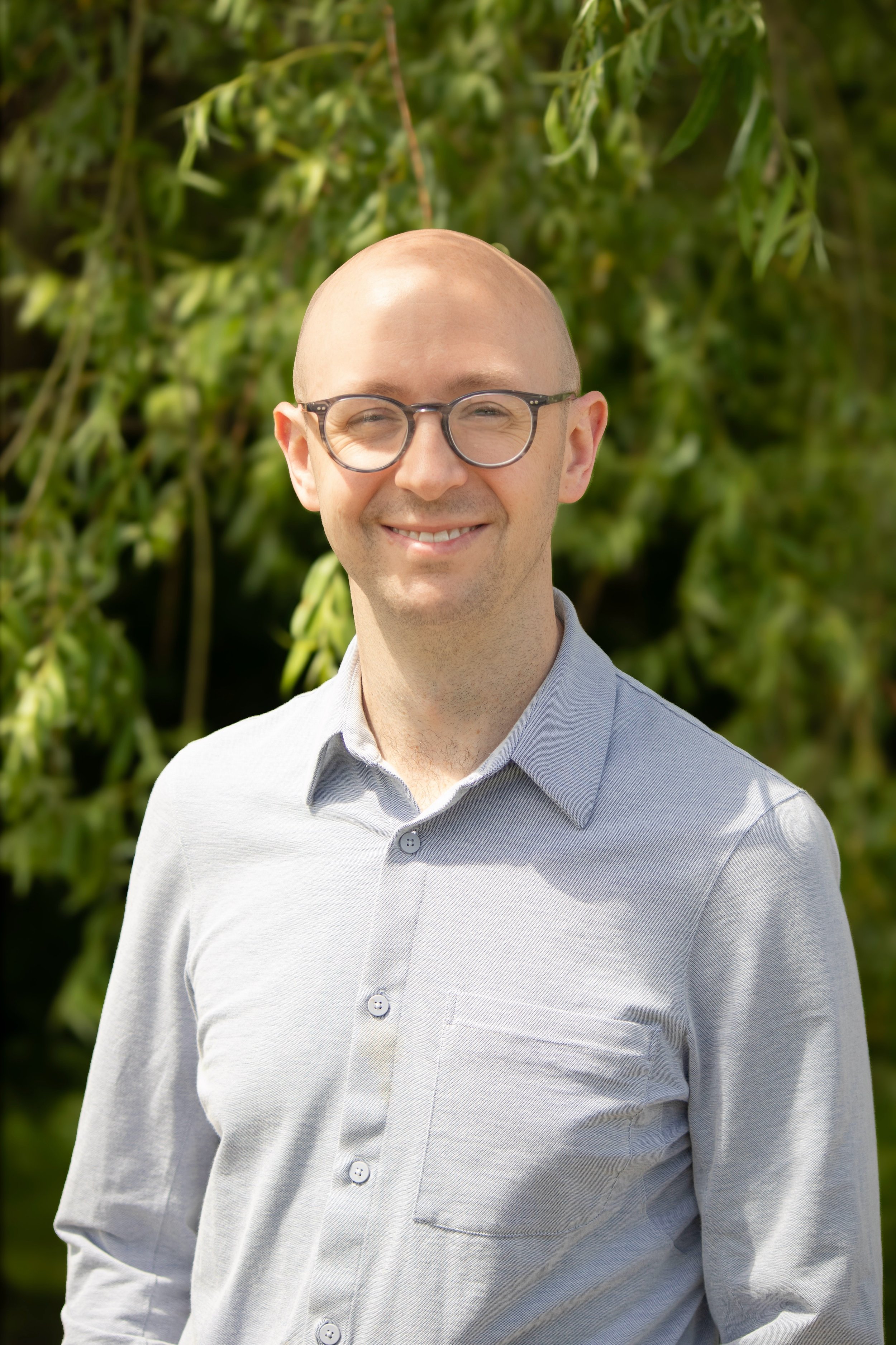 A smiling man with glasses and a bald head, wearing a light gray button-up shirt, standing outdoors with green foliage in the background.