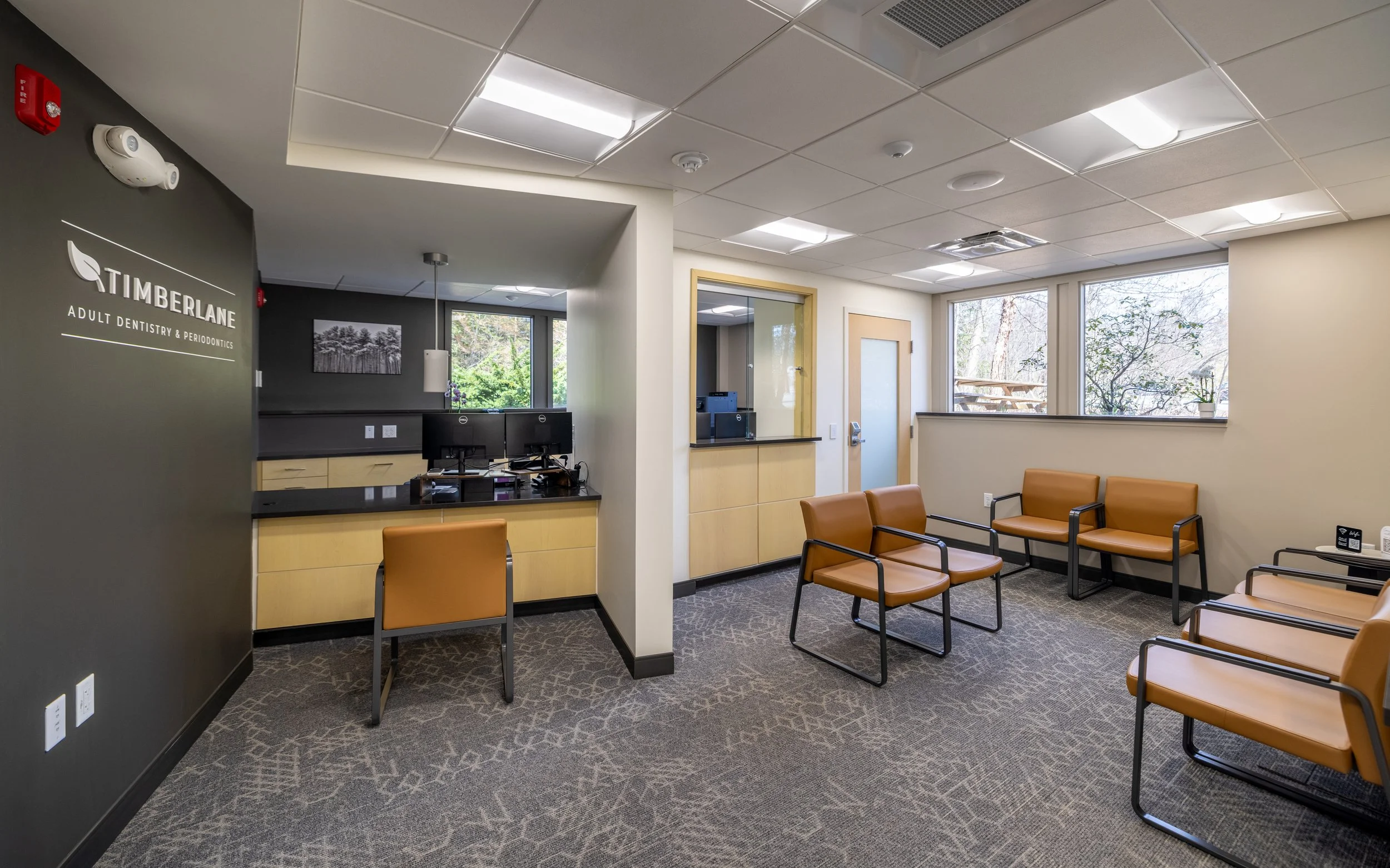 Waiting room of a dental clinic with brown chairs, a reception desk, and a window showing trees outside.