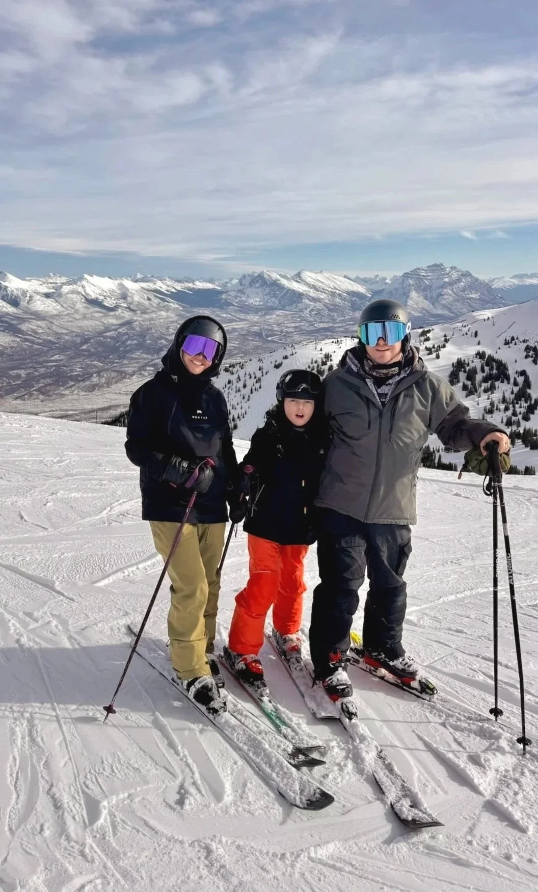 Three people skiing on snow-covered mountain with snow-capped peaks in the background, wearing winter gear and helmets.