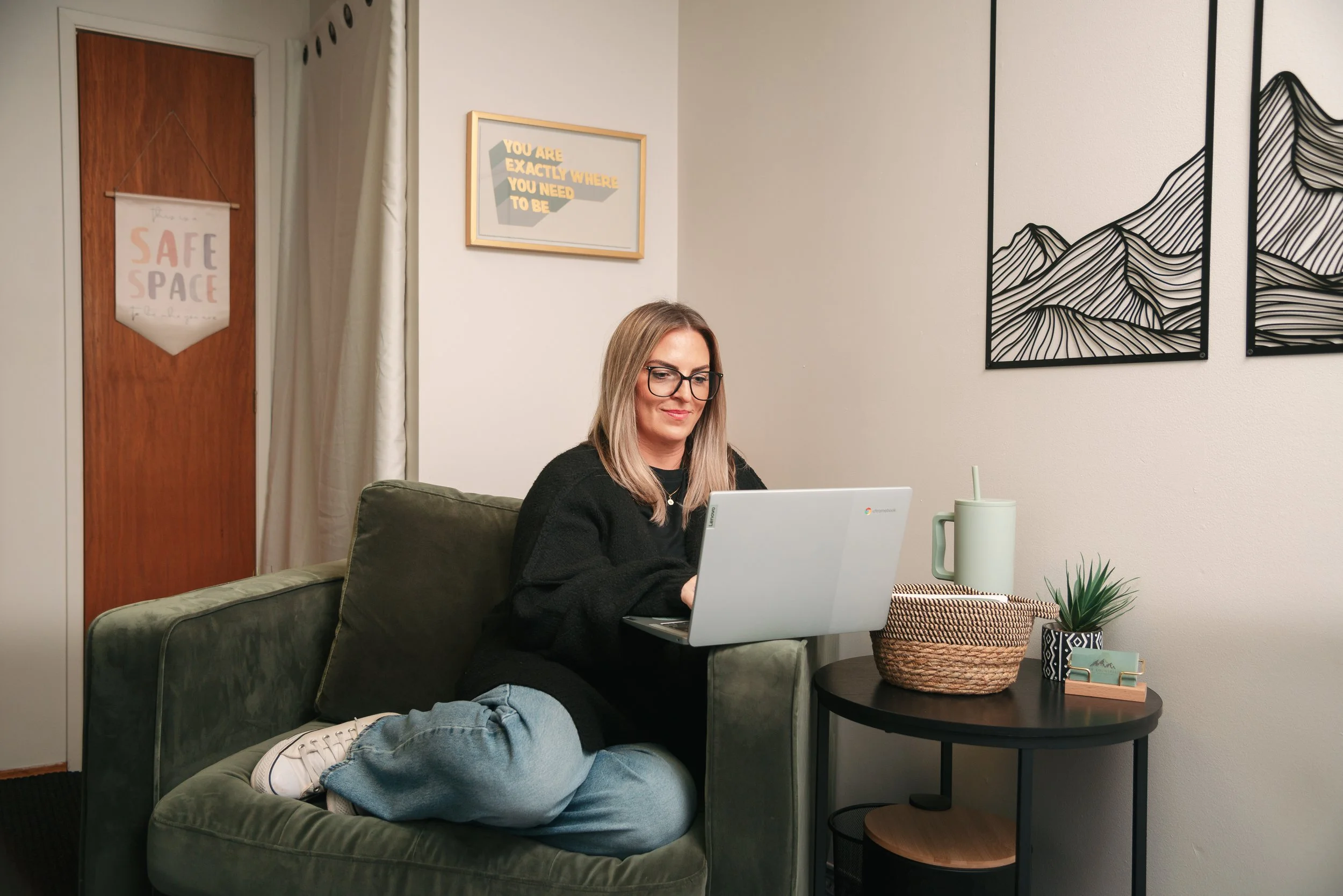 A woman with glasses sitting on a green velvet sofa, working on a white Chromebook in a cozy living room. There is a side table with a plant, a woven basket, a large mug, and a small decorative item. The wall behind has black art prints of mountain lines and inspirational framed messages. A door with a 'SAFE SPACE' banner is in the background.