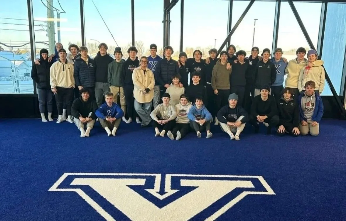 Group of young people and a woman posing indoors on a blue carpet with a large white logo, windows in the background showing a winter scene