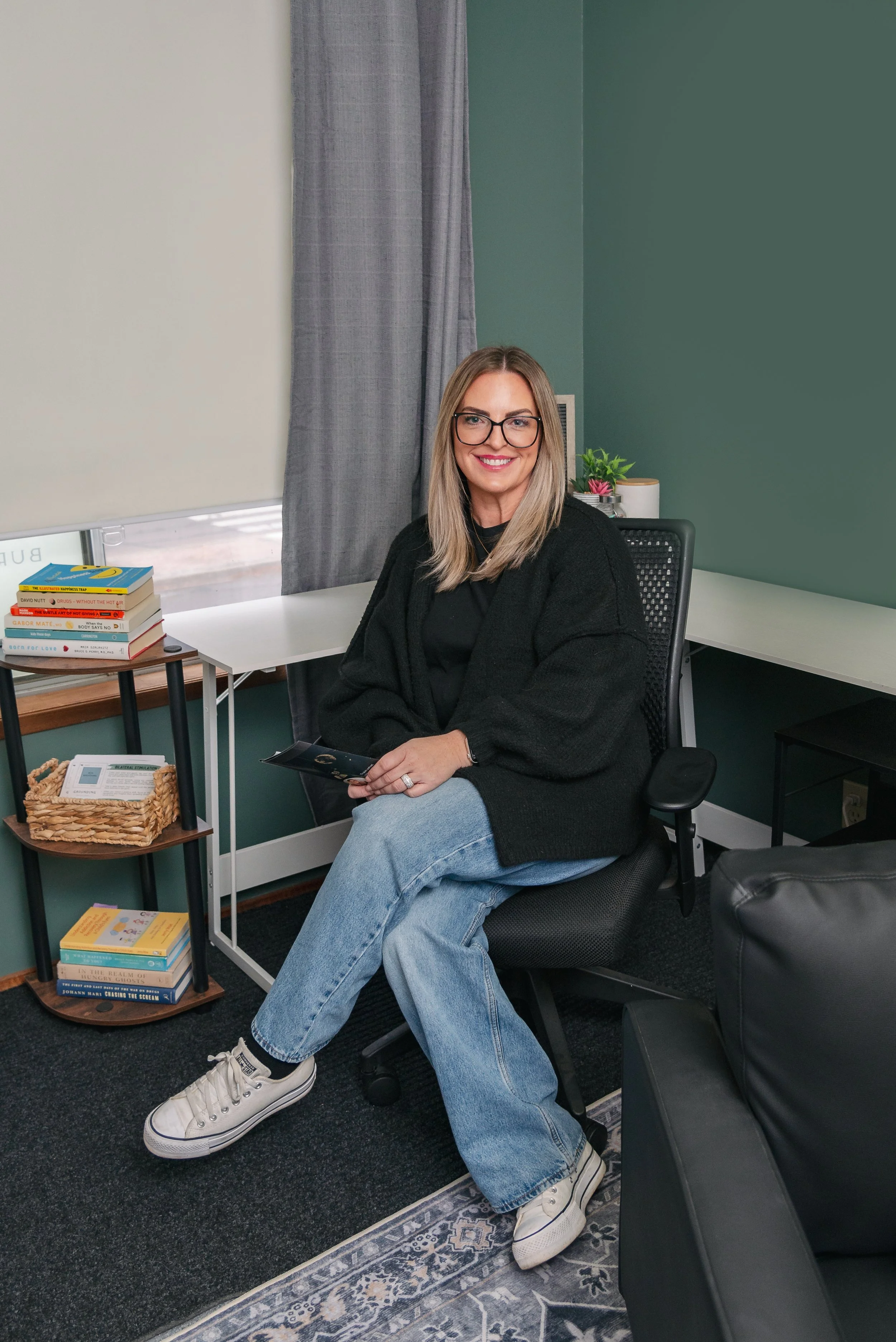 A woman with blonde hair, glasses, and a black sweater sitting on a black office chair in a room with green walls, gray curtains, and a window with blinds, holding a phone and smiling.