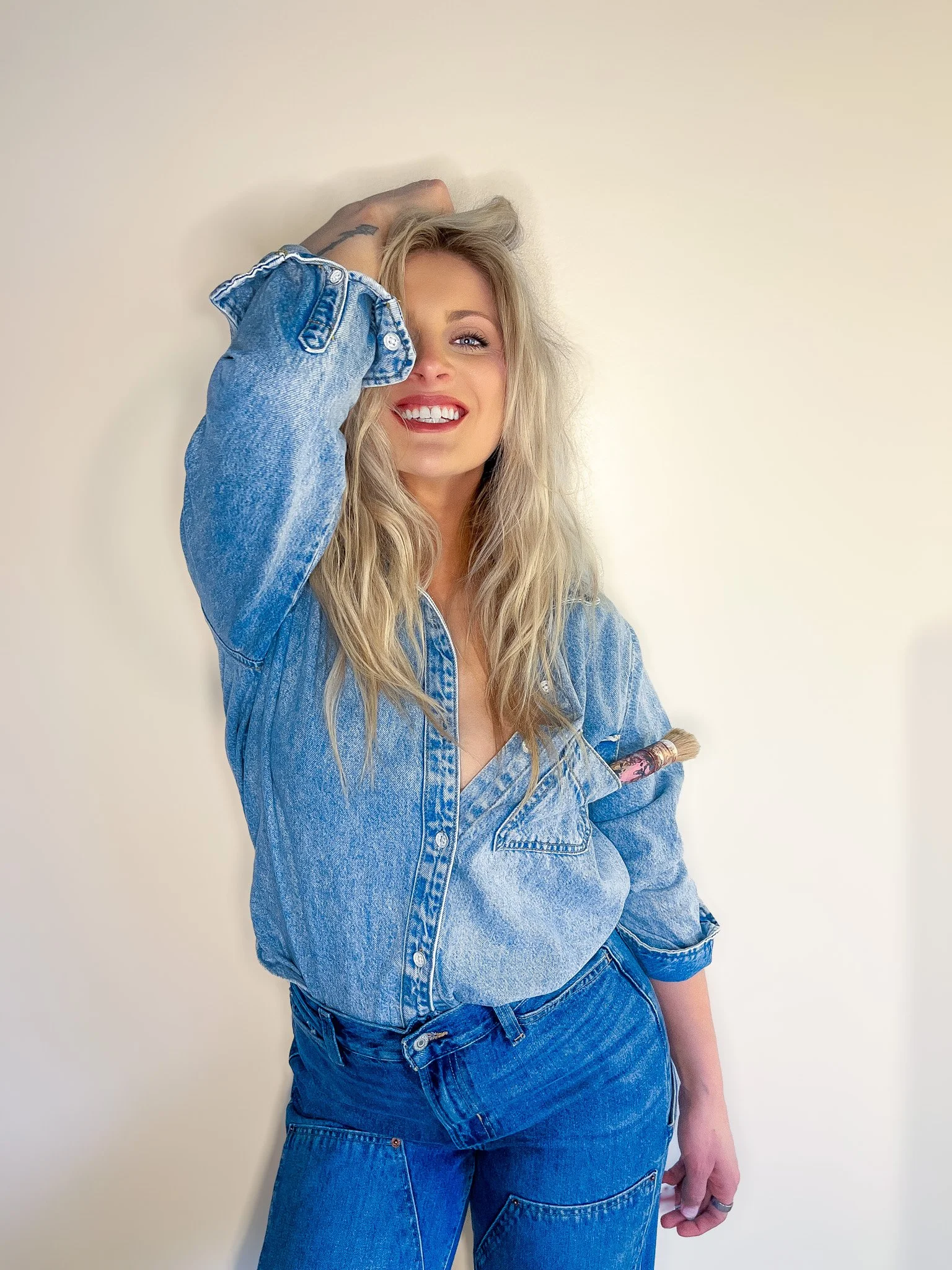 A woman with long blonde hair wearing a denim jacket and jeans, smiling and posing with one arm raised against a plain white wall.