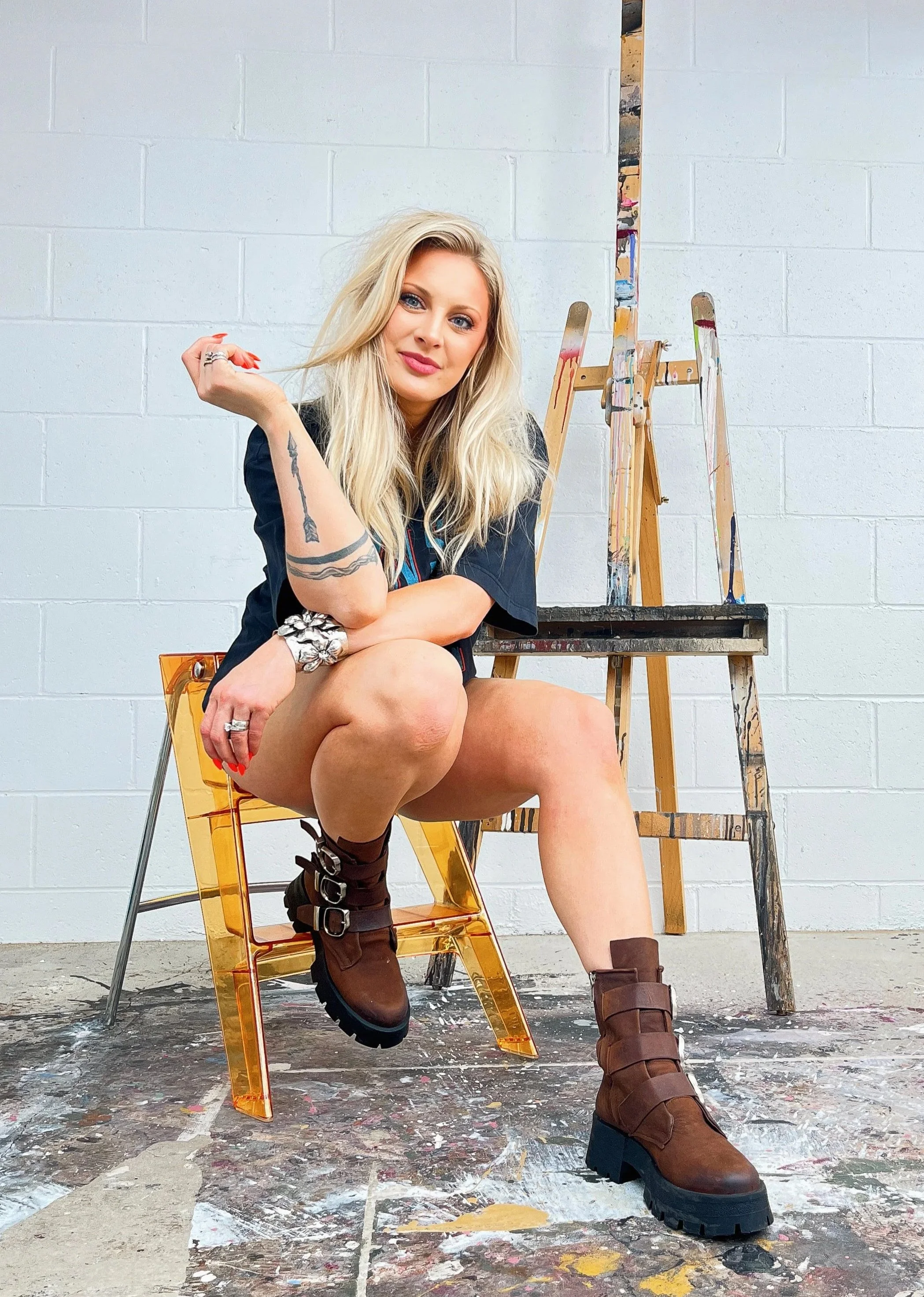 A woman with long blonde hair and tattoos is sitting on a transparent orange chair in an art studio, with a paint-splattered floor and two paint easels behind her, wearing brown boots and a black shirt.