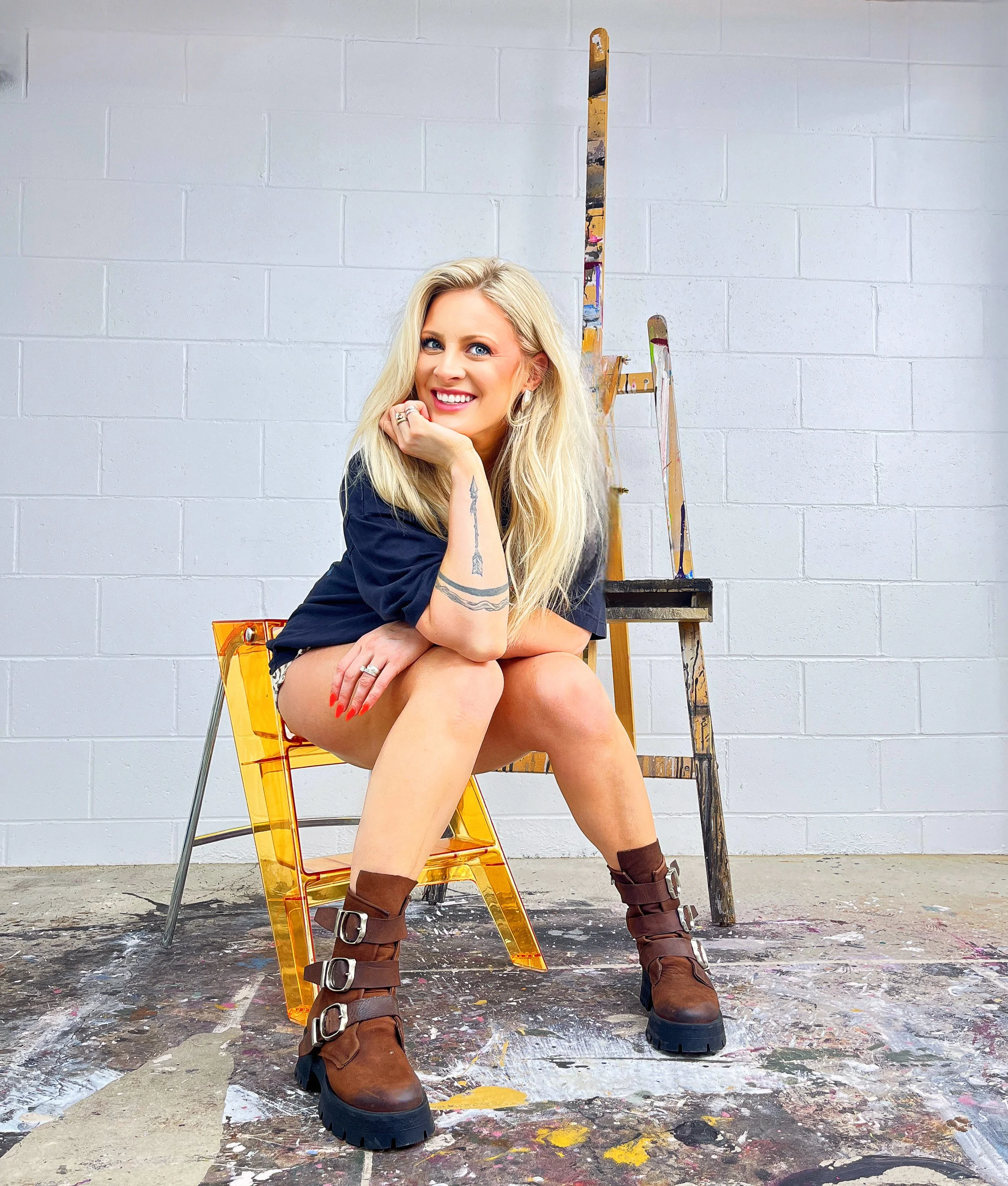 A woman with long blonde hair sitting on a yellow transparent chair in front of a gray painted cinder block wall, with an artist's palette and easel behind her, wearing brown lace-up boots and a navy shirt, smiling at the camera.