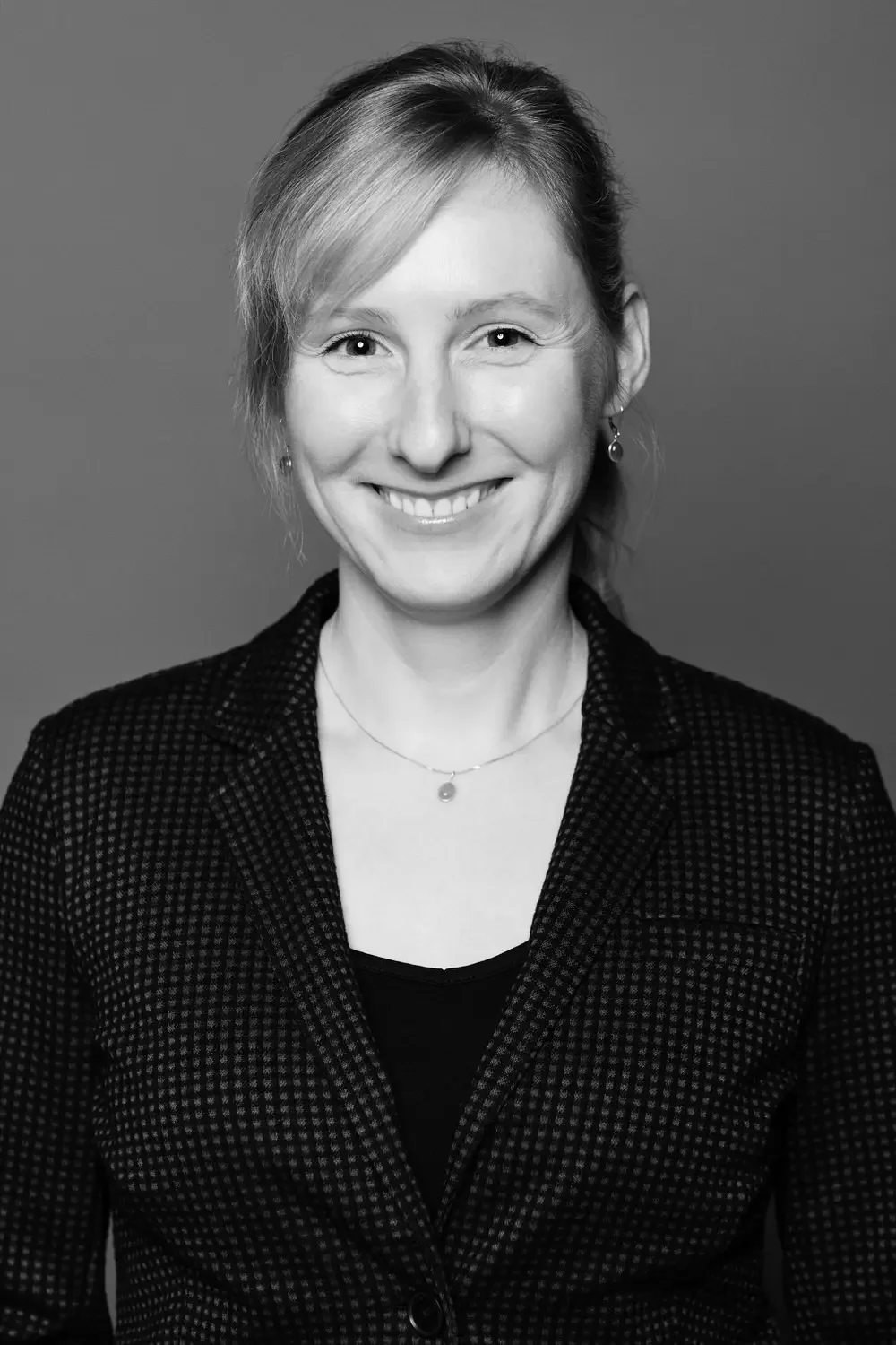 Schwarz-weiß Porträt einer lachenden Frau mit kurz gestyltem Haar, tragender schwarzer Jacke und Schmuck, vor einem neutralen Hintergrund.
