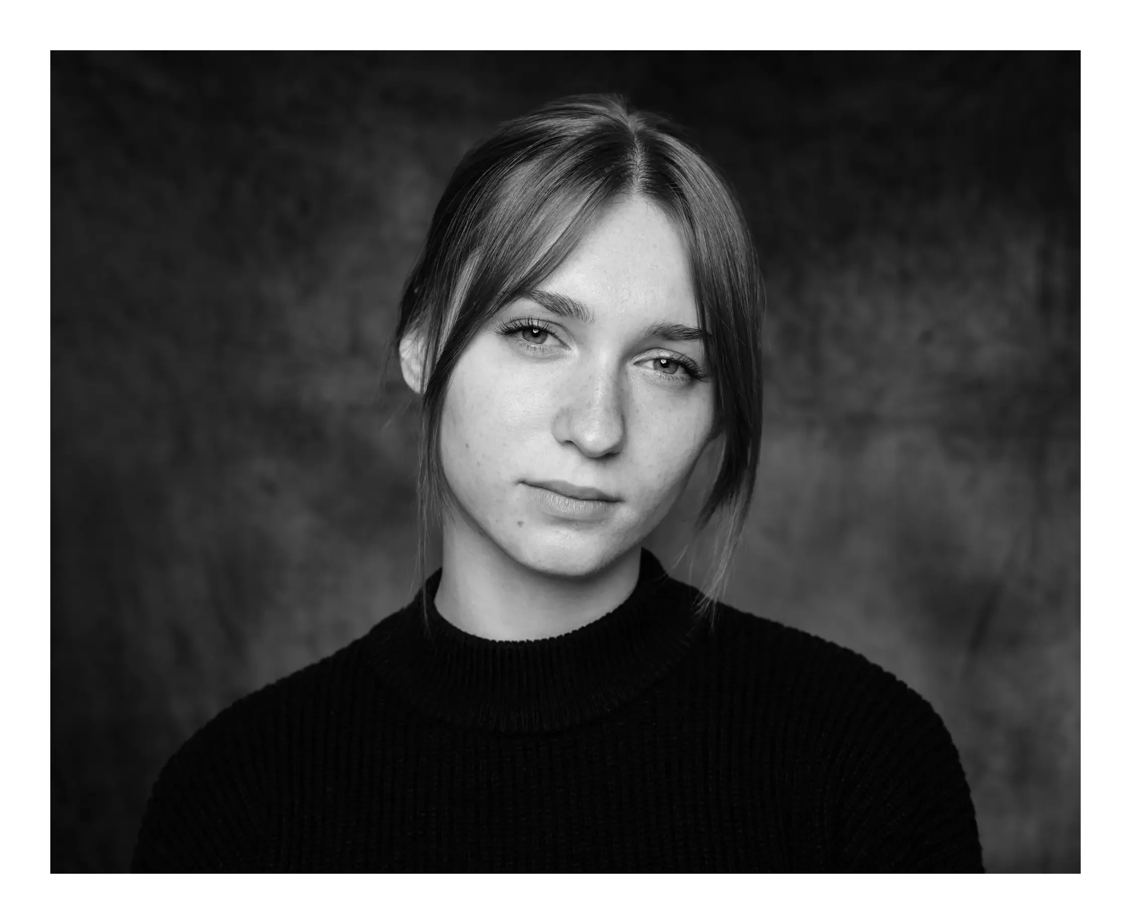 Schwarz-Weiß-Porträt einer jungen Frau mit Schulterlänge Haar, die einen schwarzen Pullover trägt, vor dunklem Hintergrund.