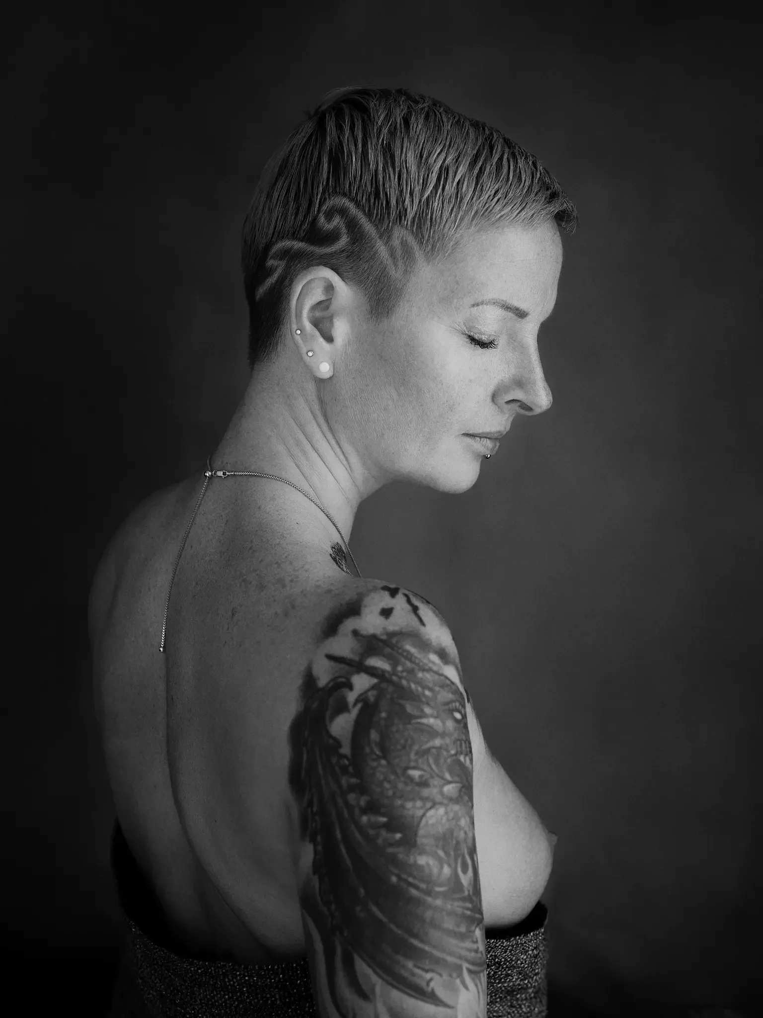 Schwarz-weiß-Fotografie einer Frau mit kurzen, seitlich geschnittenen Haaren, sichtbare Tätowierung am Oberarm, trägt Ohrringe und eine Halskette, blickt nach unten, neutraler Hintergrund.