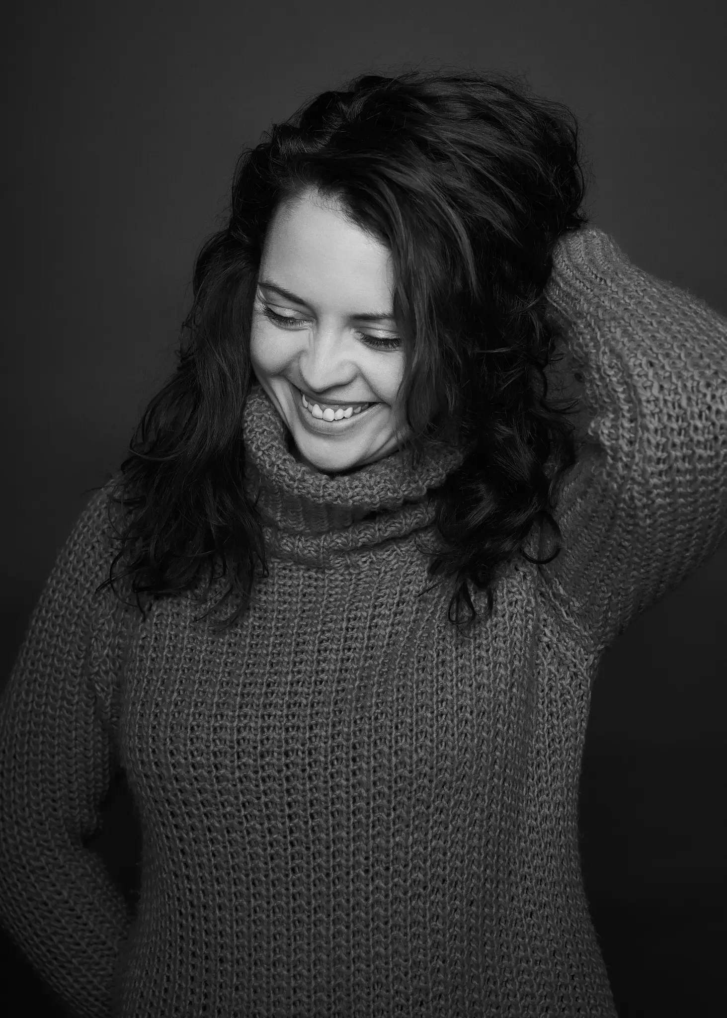 Schwarz-Weiß-Foto einer lachenden Frau, die ein gestricktes Tuch trägt und ihre Hand im Haar hat.