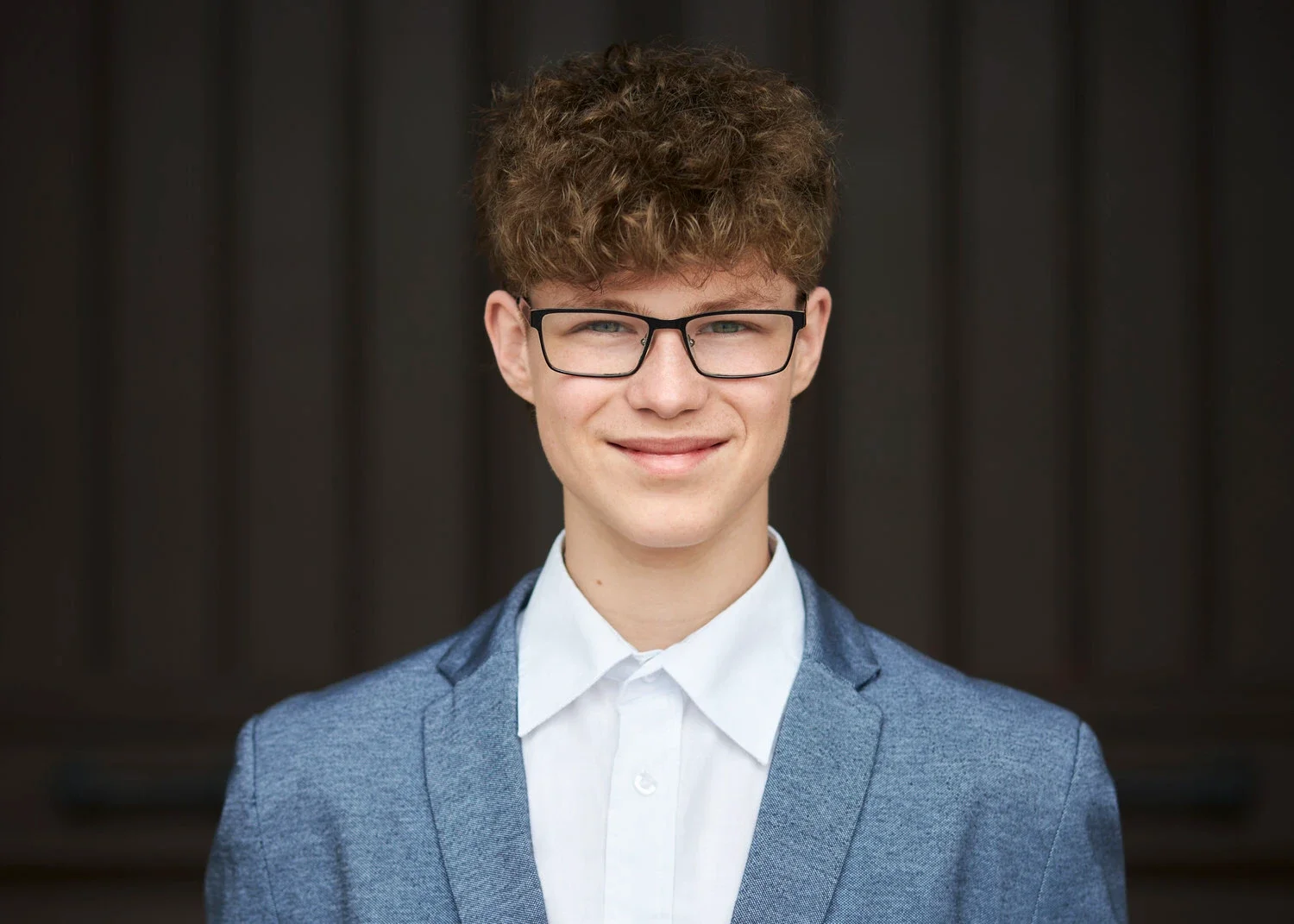 Porträt eines jungen Mannes in einem blauen Anzug und weißen Hemd, mit Brille, vor dunklem Hintergrund, lächelnd.