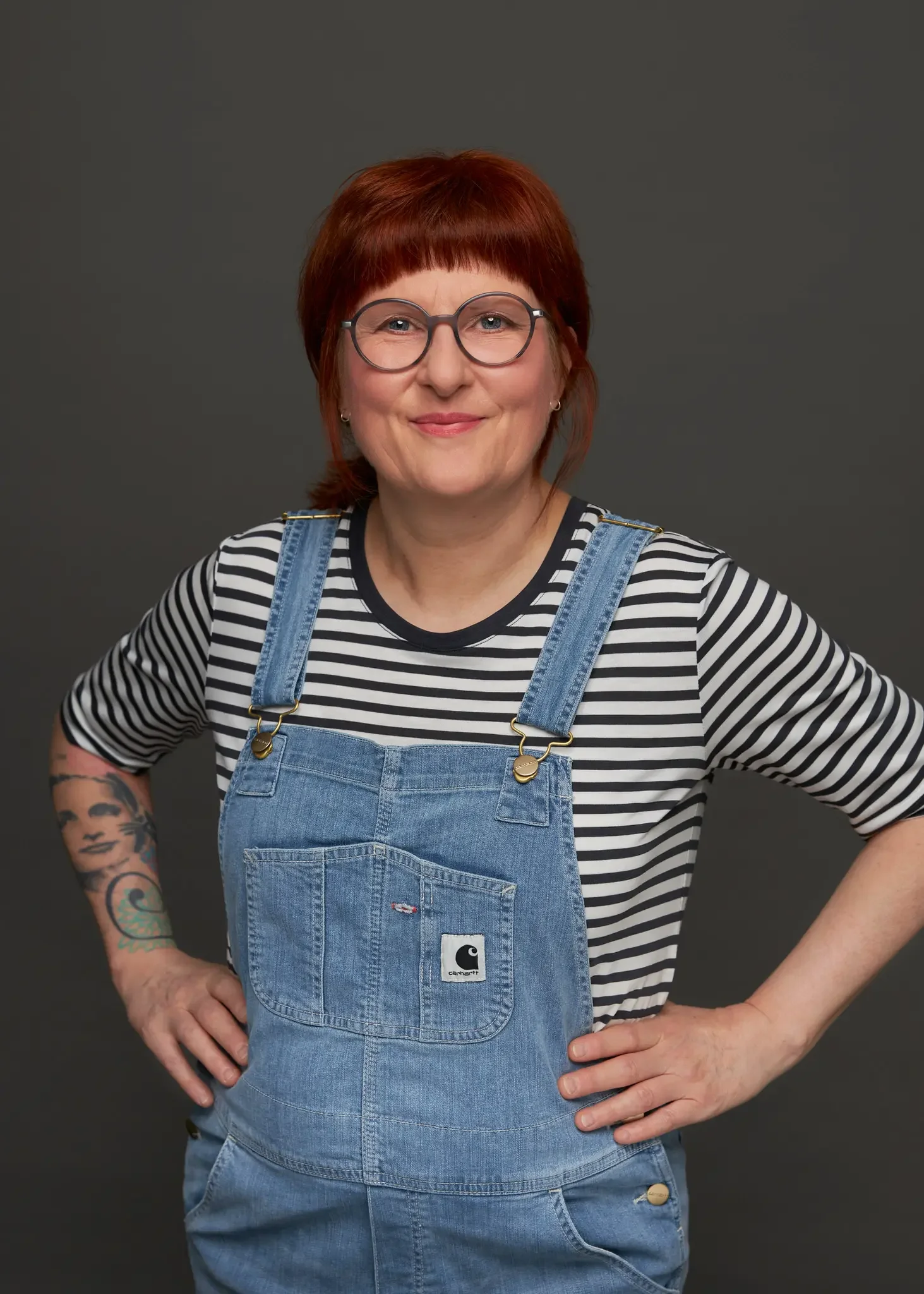 Frau mit roten Haaren, Brille, gestreiftem T-Shirt und Jeans-Overall, steht vor grauem Hintergrund.